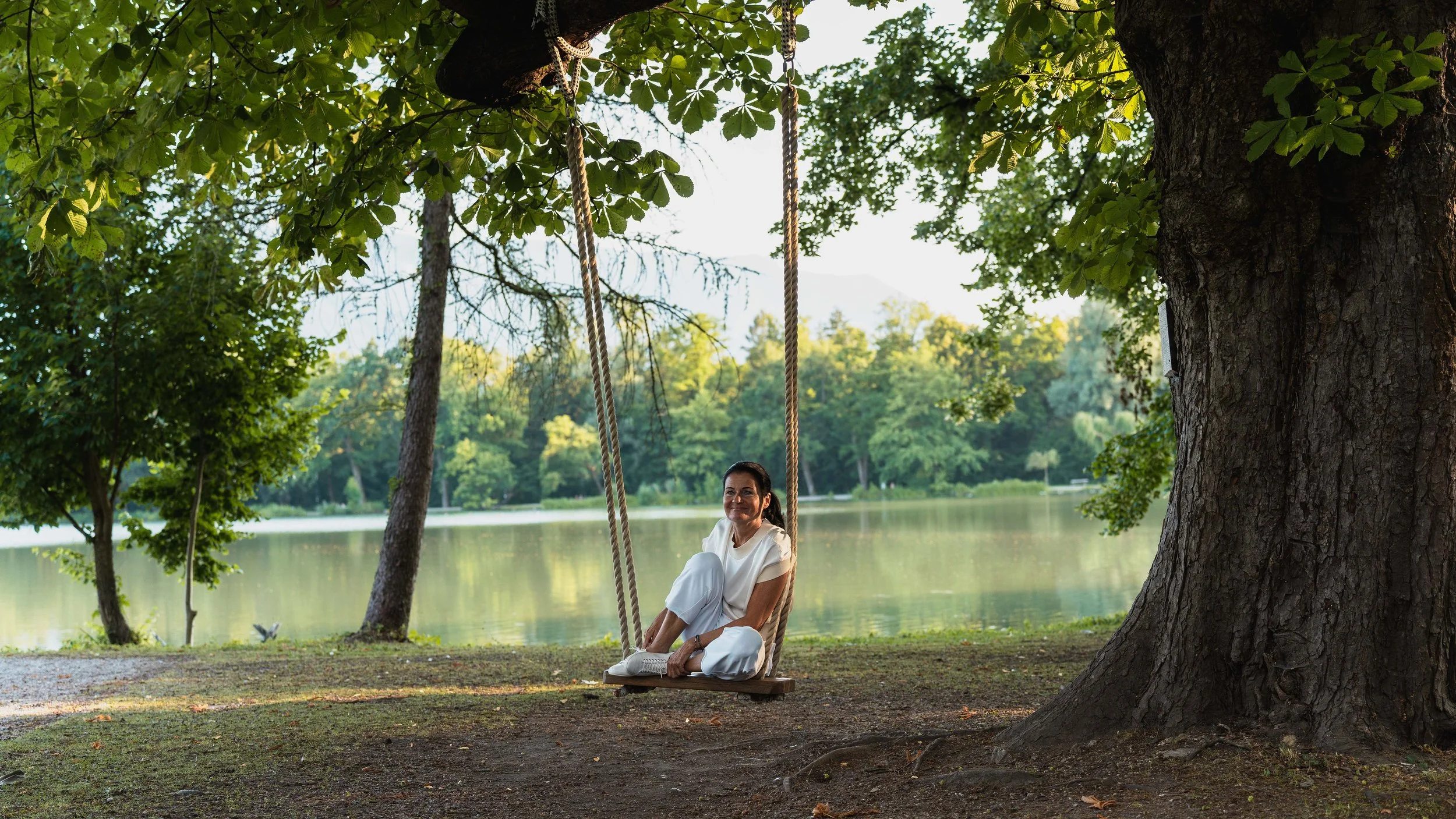 Eine Frau sitzt entspannt auf einer Schaukel unter einem großen Baum am Seeufer, umgeben von grünen Blättern und Bäumen, mit Wasser im Hintergrund und einem bewölkten Himmel.