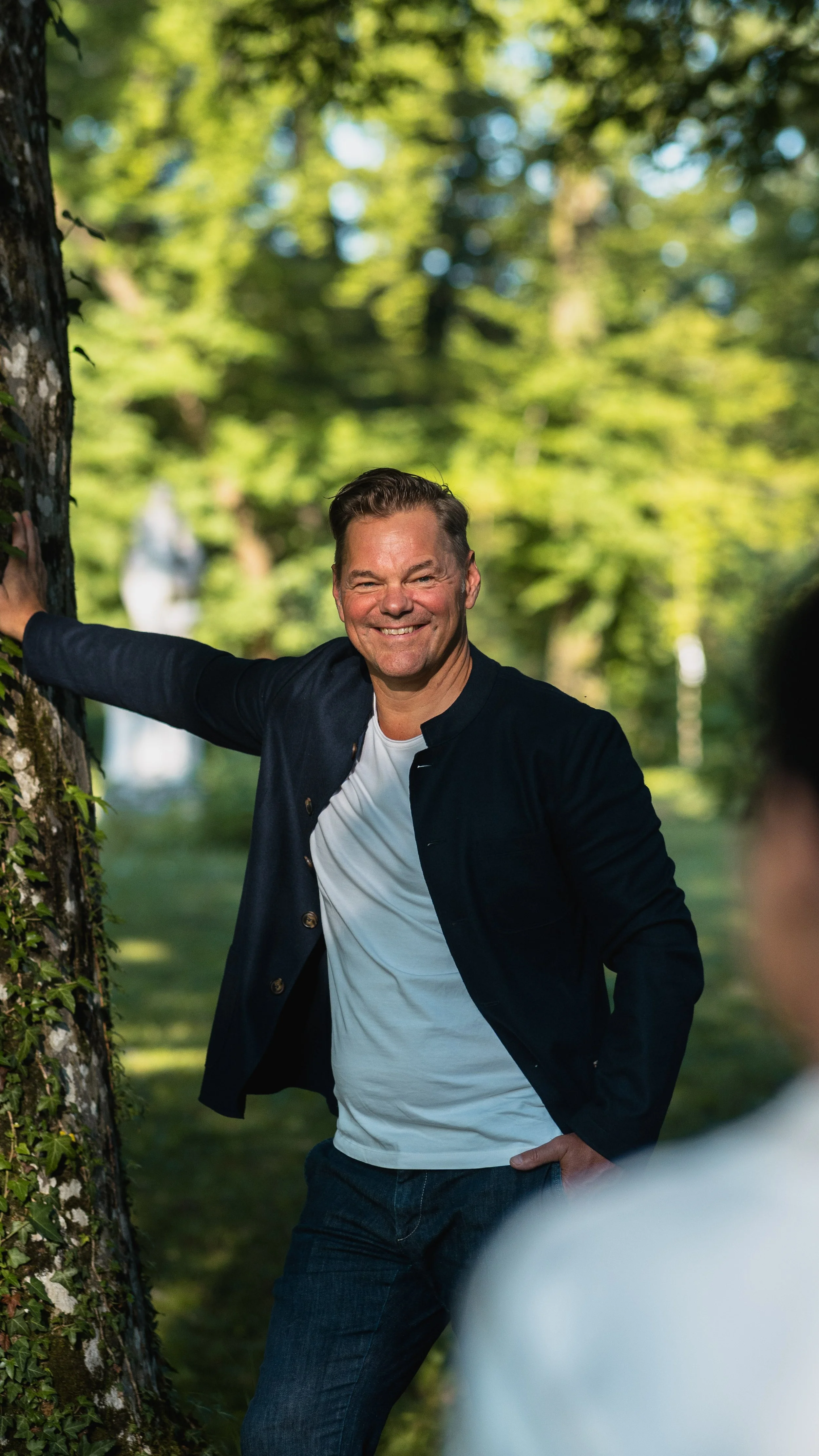 Ein lachender Mann steht im Park neben einem Baum und posiert für die Kamera, mit einem blauen Himmel im Hintergrund.