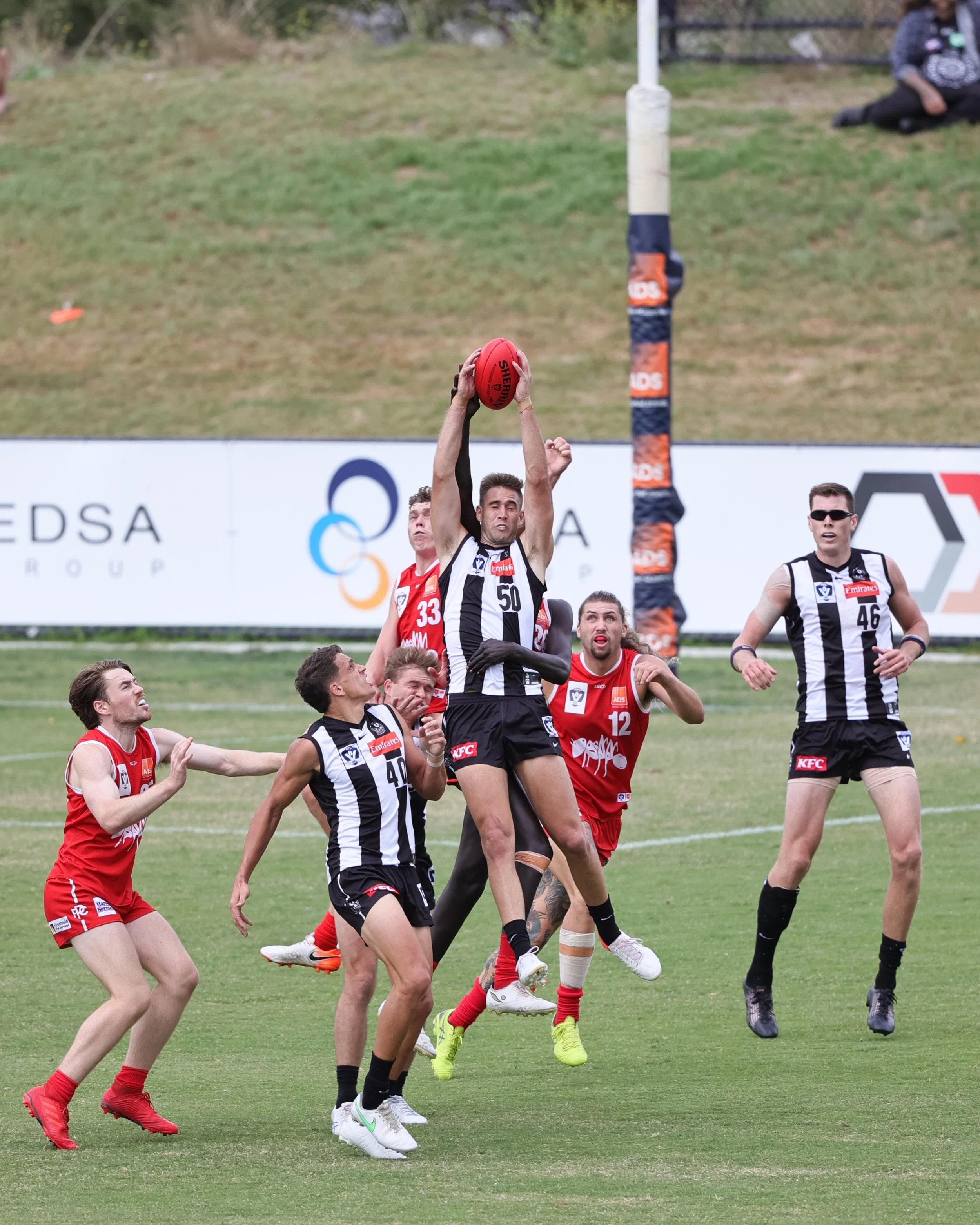 A group of Australian rules football players in black and white striped and red uniforms compete for a mark near the goal post on a grassy field.