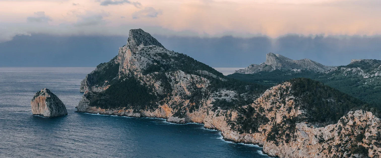 Rocky island formations with a large cliff in the background, surrounded by the ocean and some clouds in the sky.