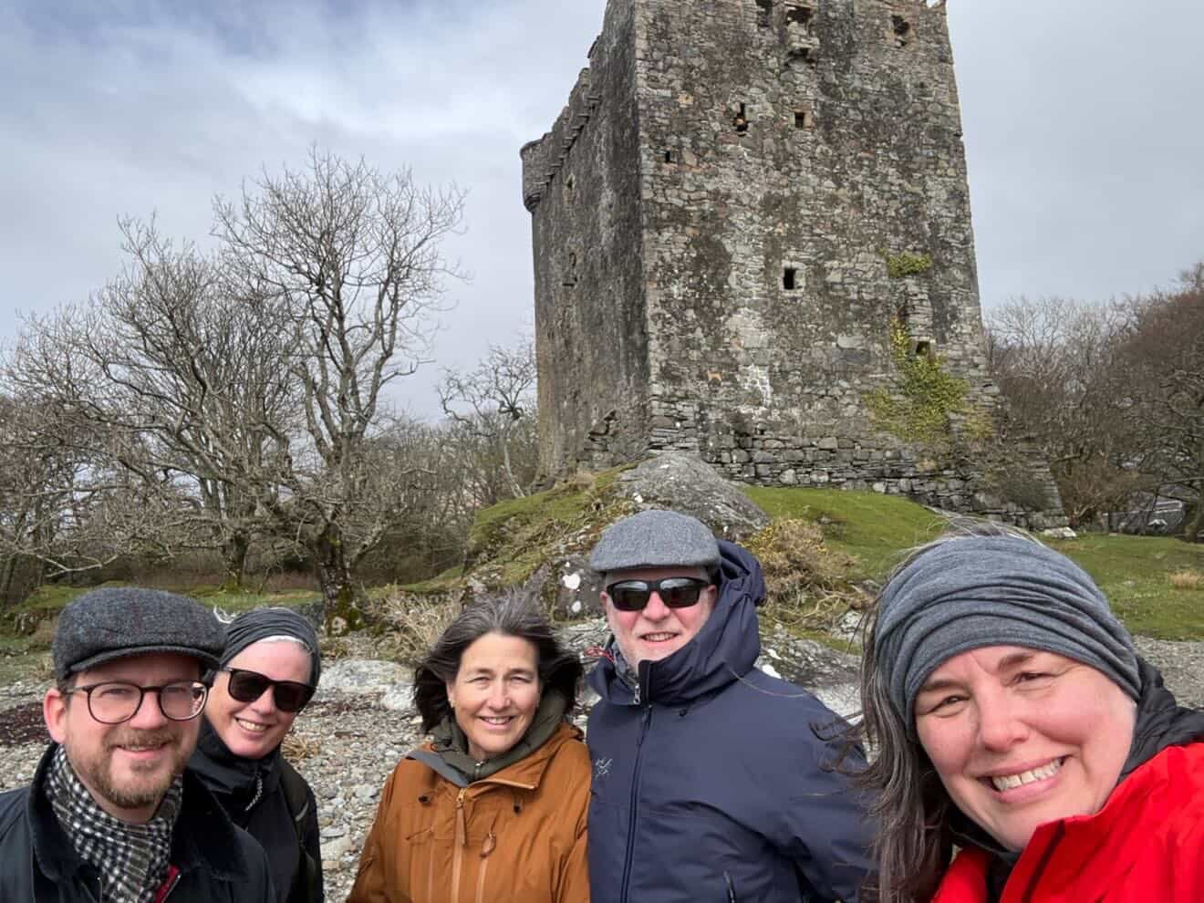 Five people smiling outdoors in front of an old stone tower, wearing jackets and hats, with leafless trees in the background on a cloudy day.