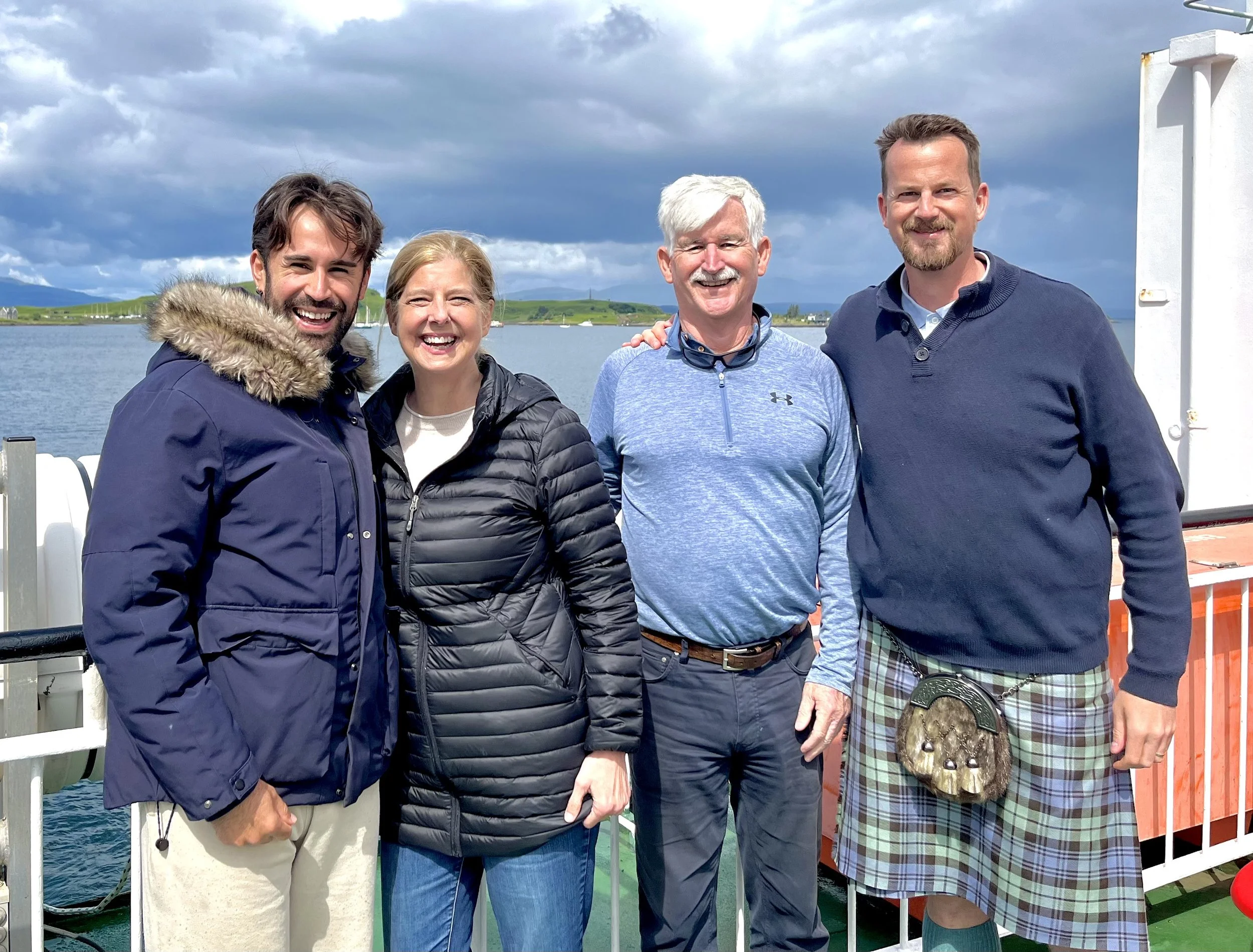 Four people standing on a ship, smiling, with a body of water and green hills in the background.