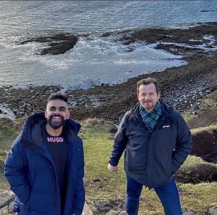 Two men smiling outdoors on grass, with a rocky shoreline and water in the background.