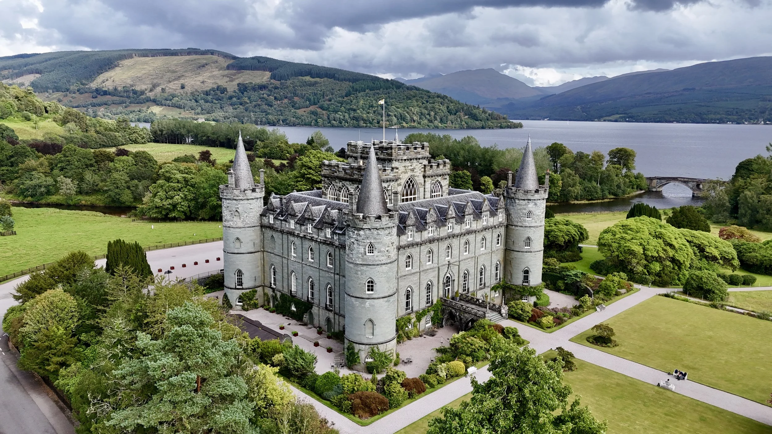 A large historic castle surrounded by lush green trees and gardens, situated near a body of water with hills and a bridge in the background.