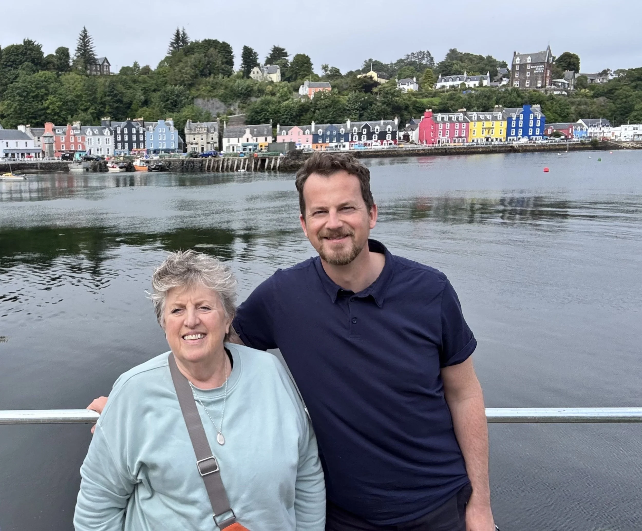 A smiling older woman and a smiling younger man pose together on a boat with a colorful riverside town and green hills in the background.