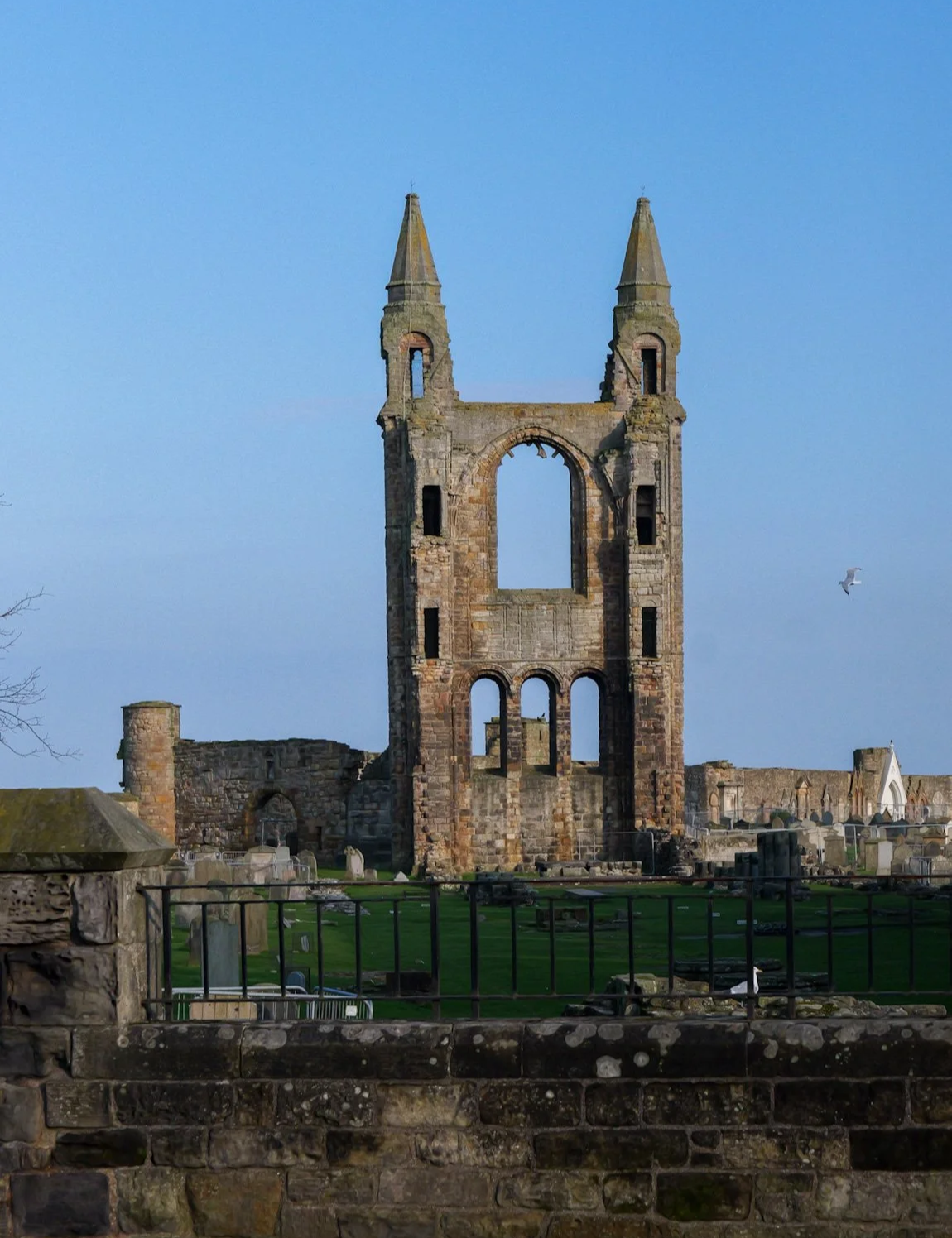An old stone church ruin with two tall steeples and a large arched window in the center, surrounded by a graveyard, under a clear blue sky with a passing bird.