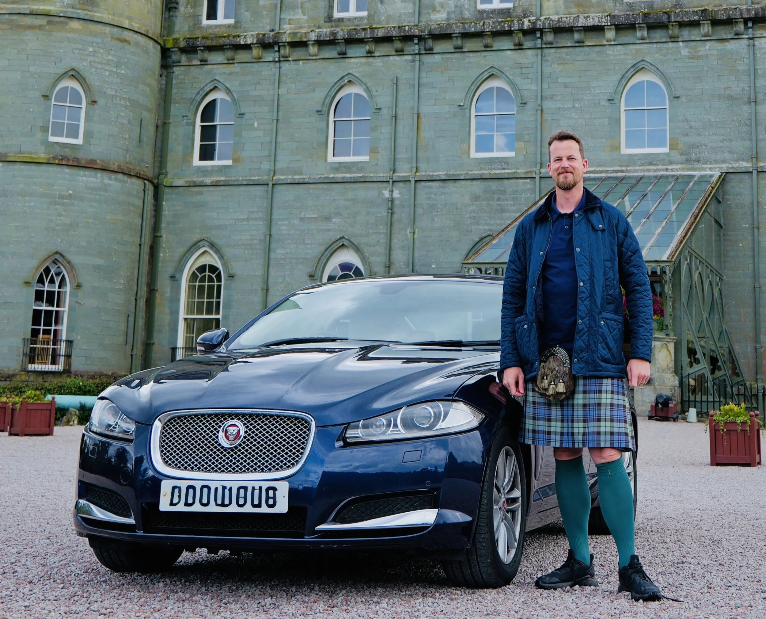 A man wearing a traditional Scottish kilt, socks, and jacket standing next to a black Jaguar car in front of an old stone building with arched windows.