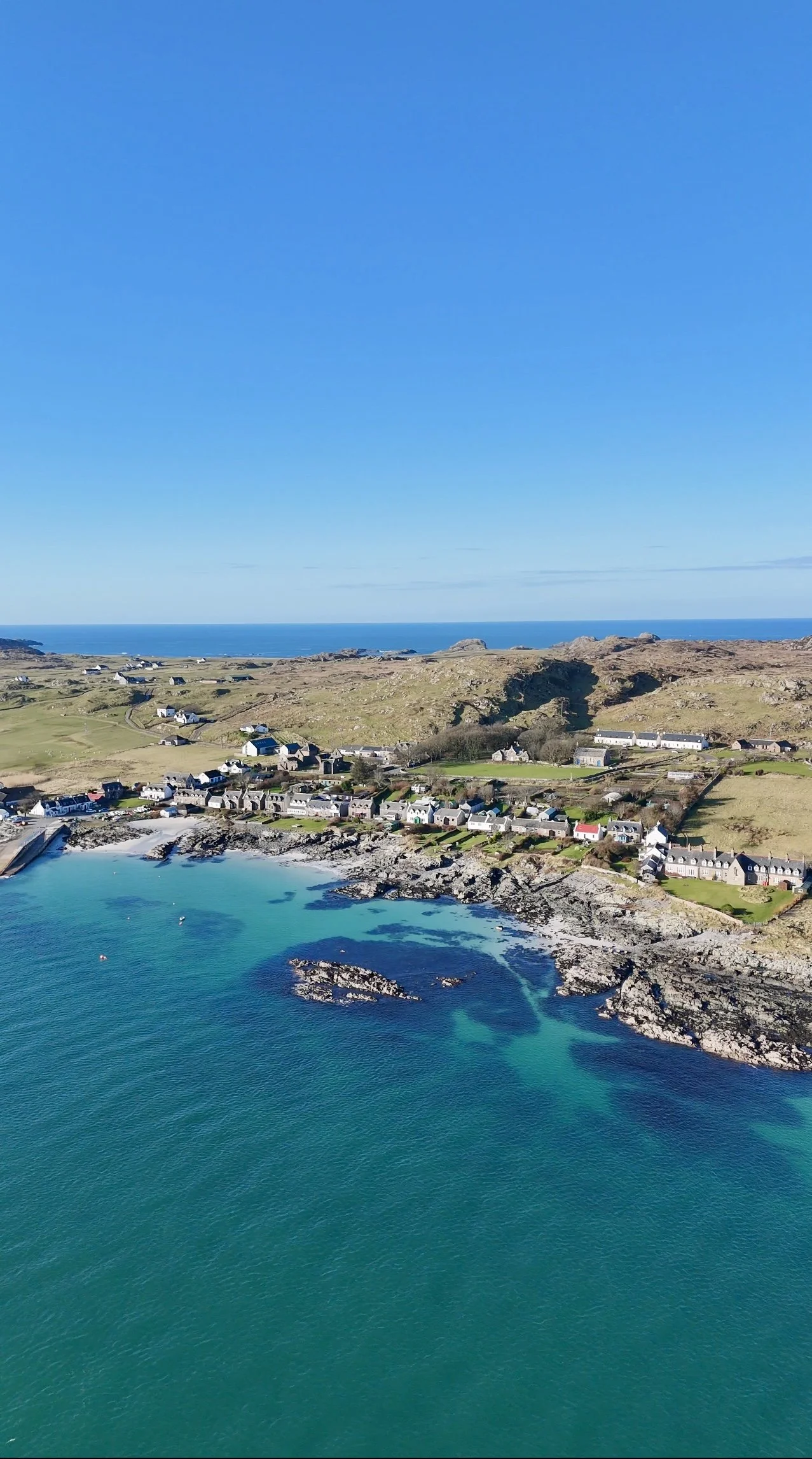 Aerial view of a coastal village with houses along the shoreline, rocky islands in the turquoise water, grassy hills, and a blue sky.