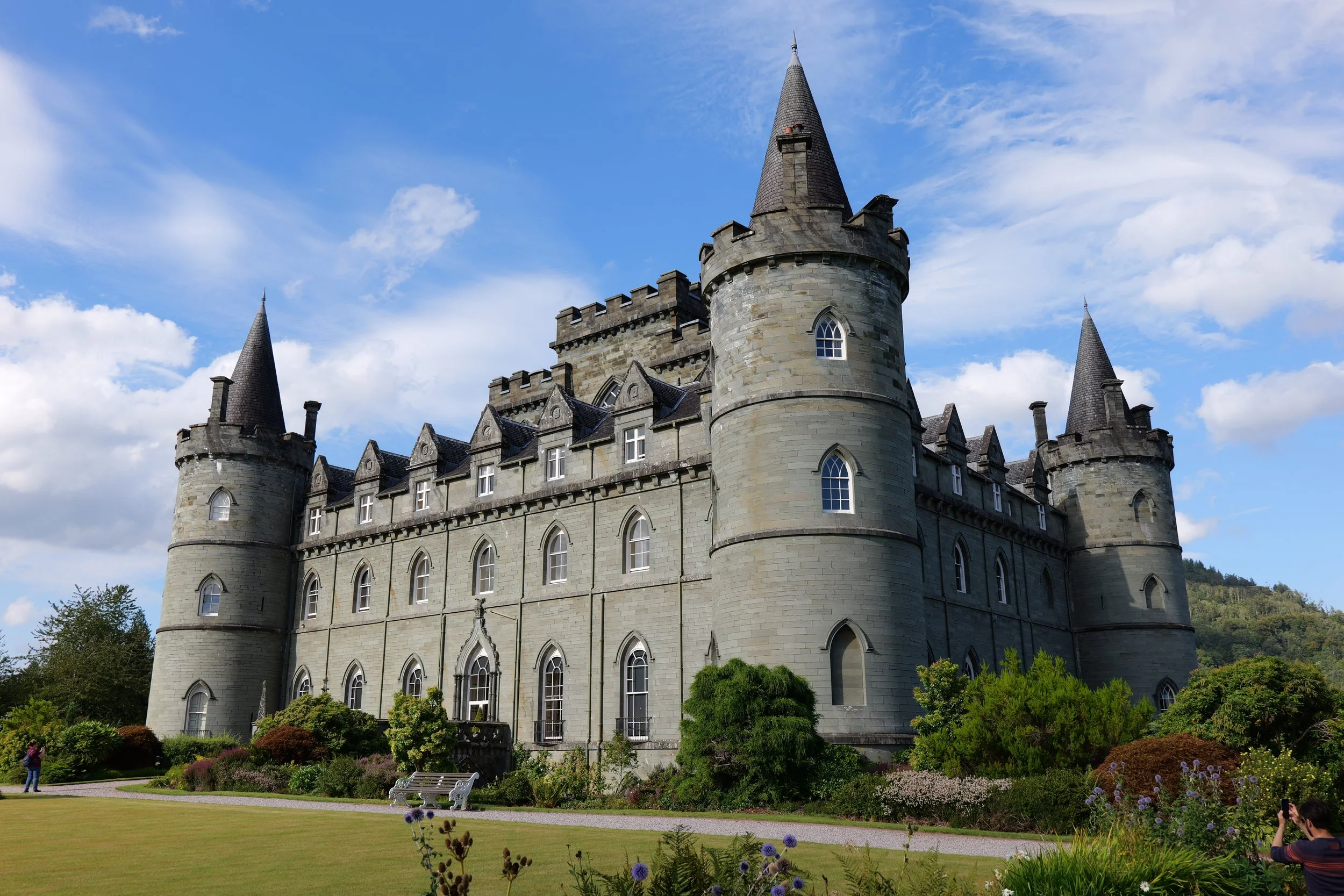 A large stone castle with multiple towers and pointed rooftops, surrounded by green bushes and plants, under a partly cloudy blue sky.