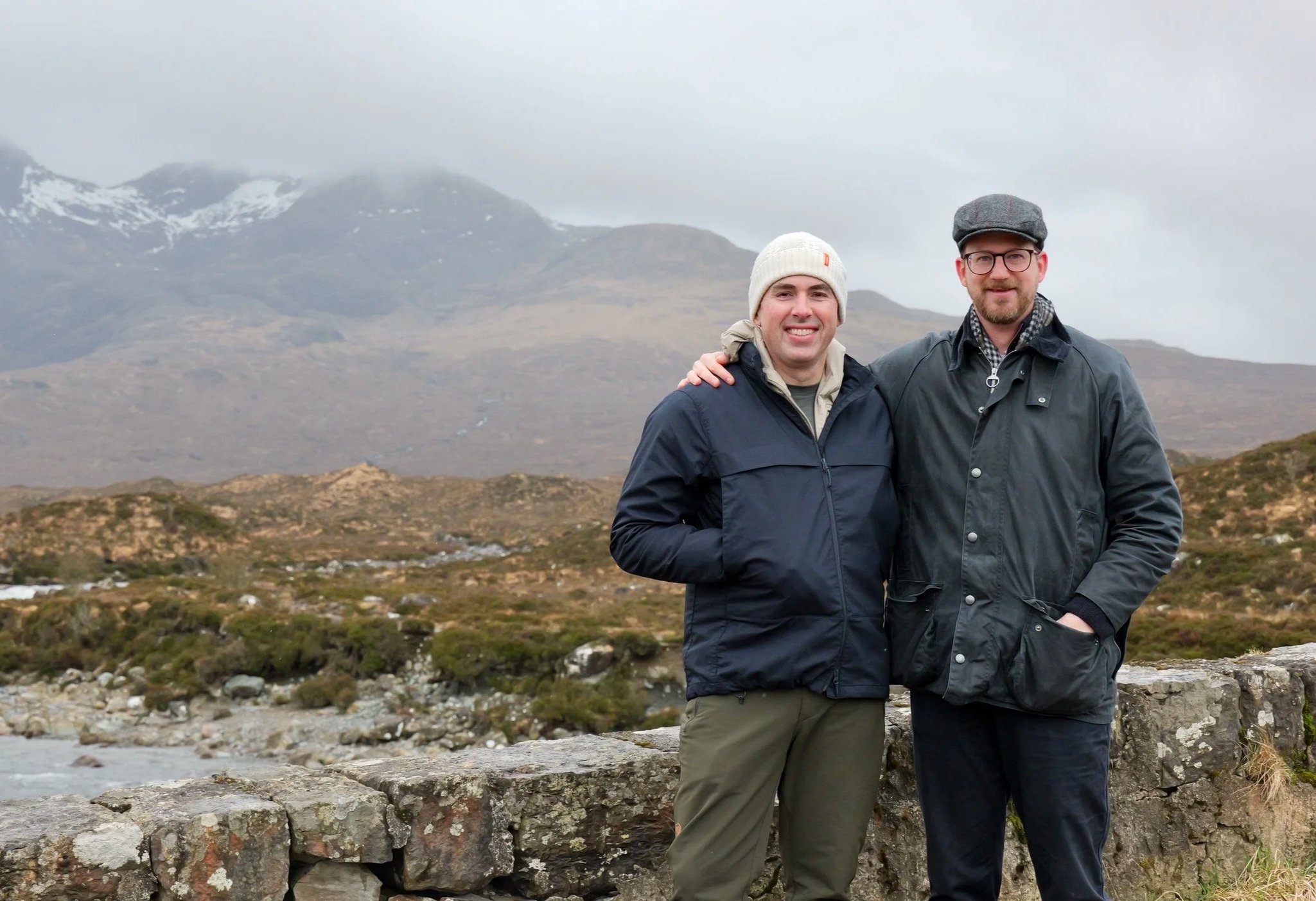 Two men standing close together outdoors in a remote mountainous landscape with cloudy sky, wearing jackets and beanies or hats, smiling