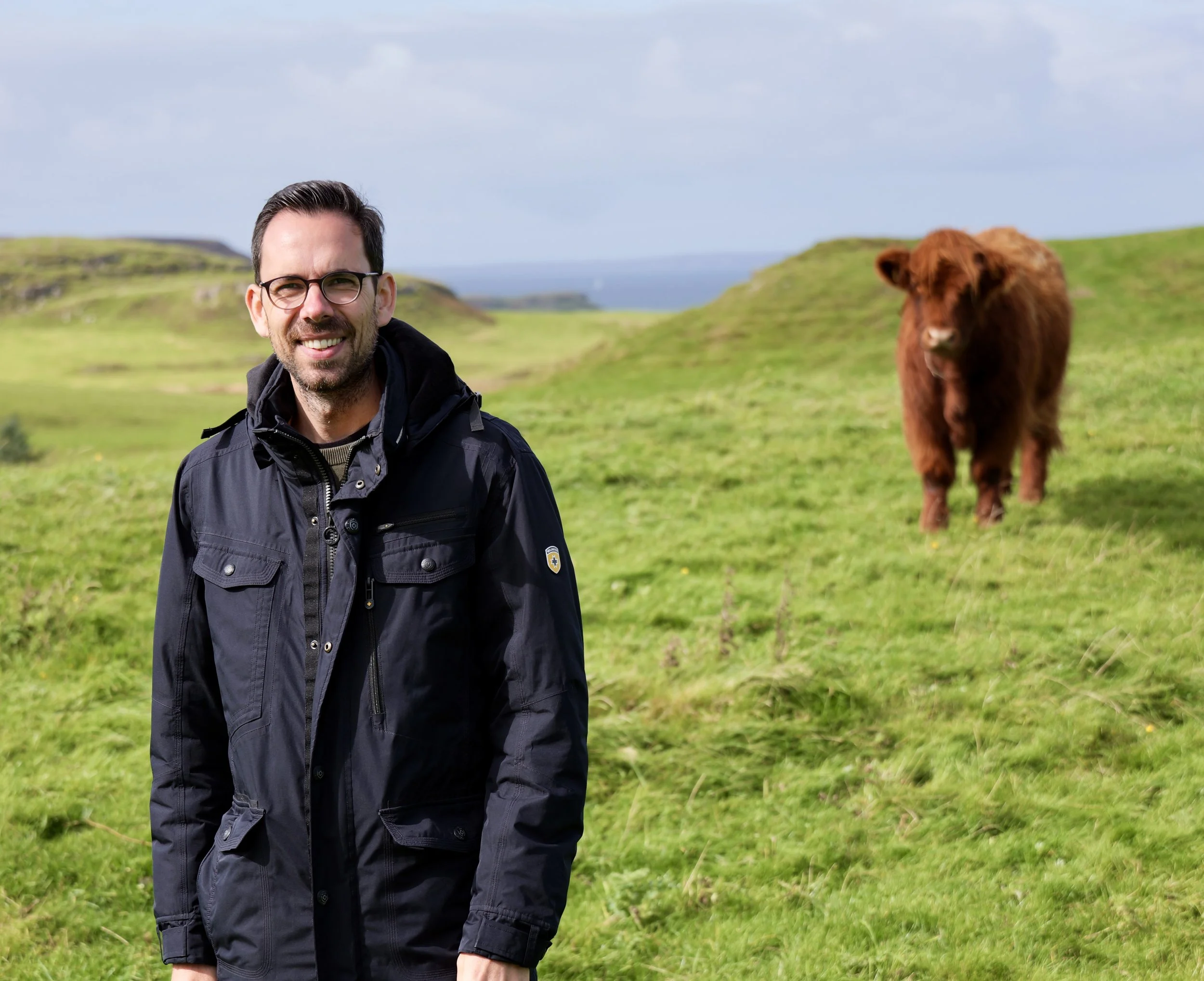 A man wearing glasses and a black jacket smiling on a green grassy hillside with a brown cow in the background.