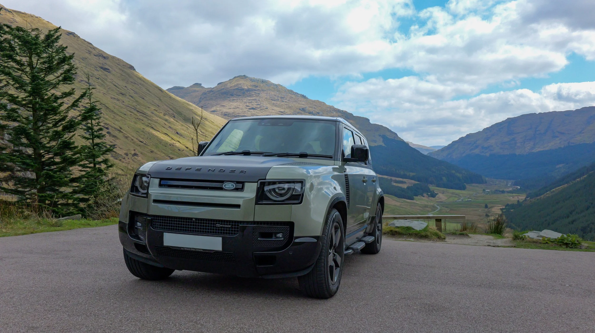 A silver Land Rover Defender parked on a mountain road with green hills and mountains in the background under a partly cloudy sky.