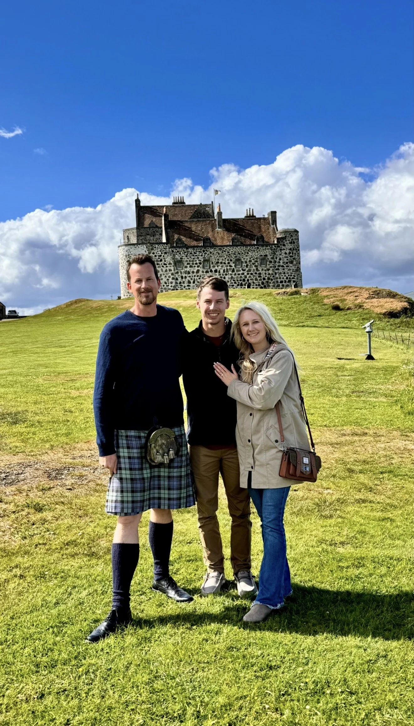 Three people standing on a grassy hill with a medieval castle in the background under a partly cloudy sky.