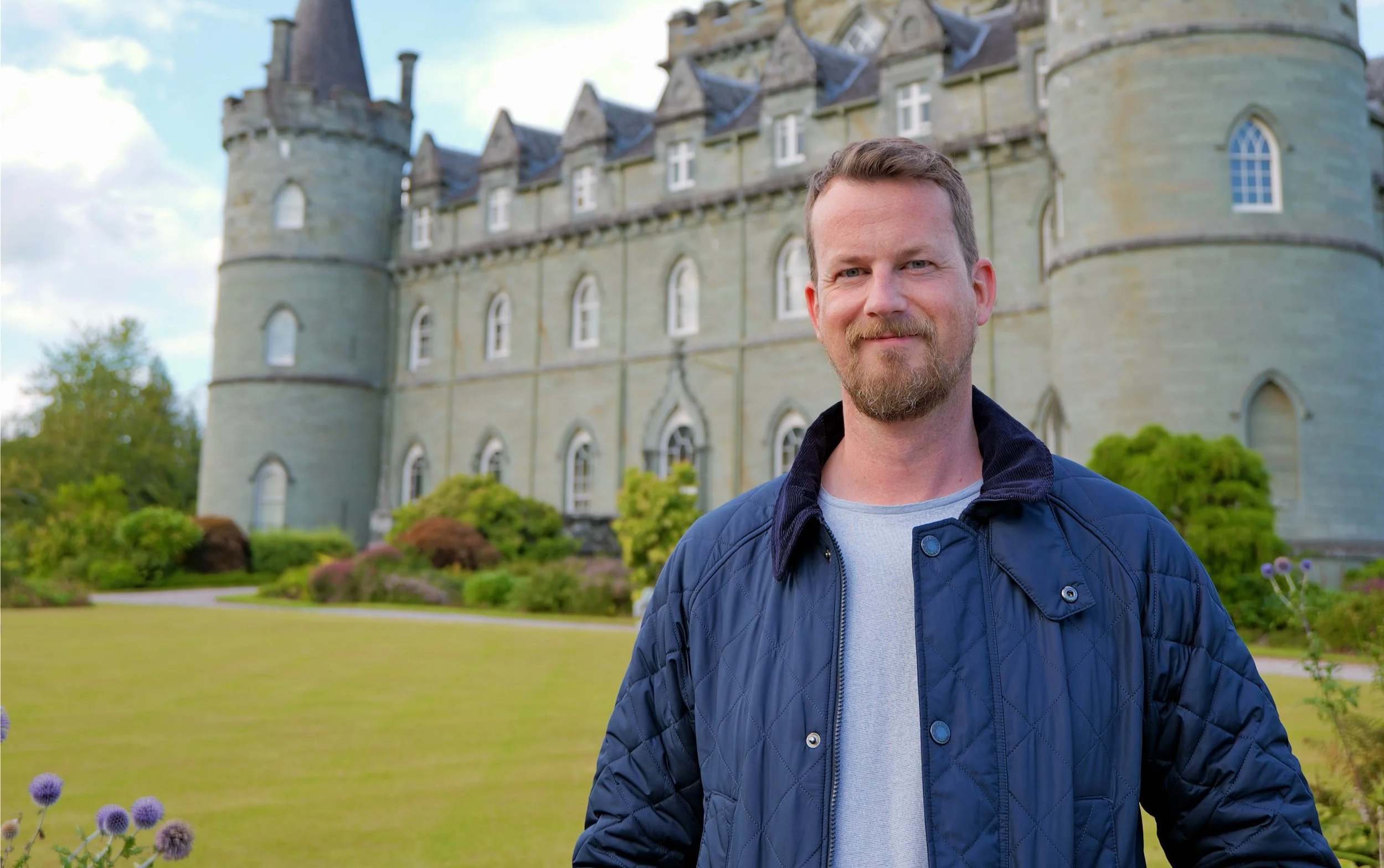 A man with a beard and short hair is standing outdoors in front of a castle with multiple turrets and large windows. He is wearing a navy quilted jacket over a light gray shirt. The background includes well-maintained grass, flowering plants, and trees. The sky is partly cloudy.
