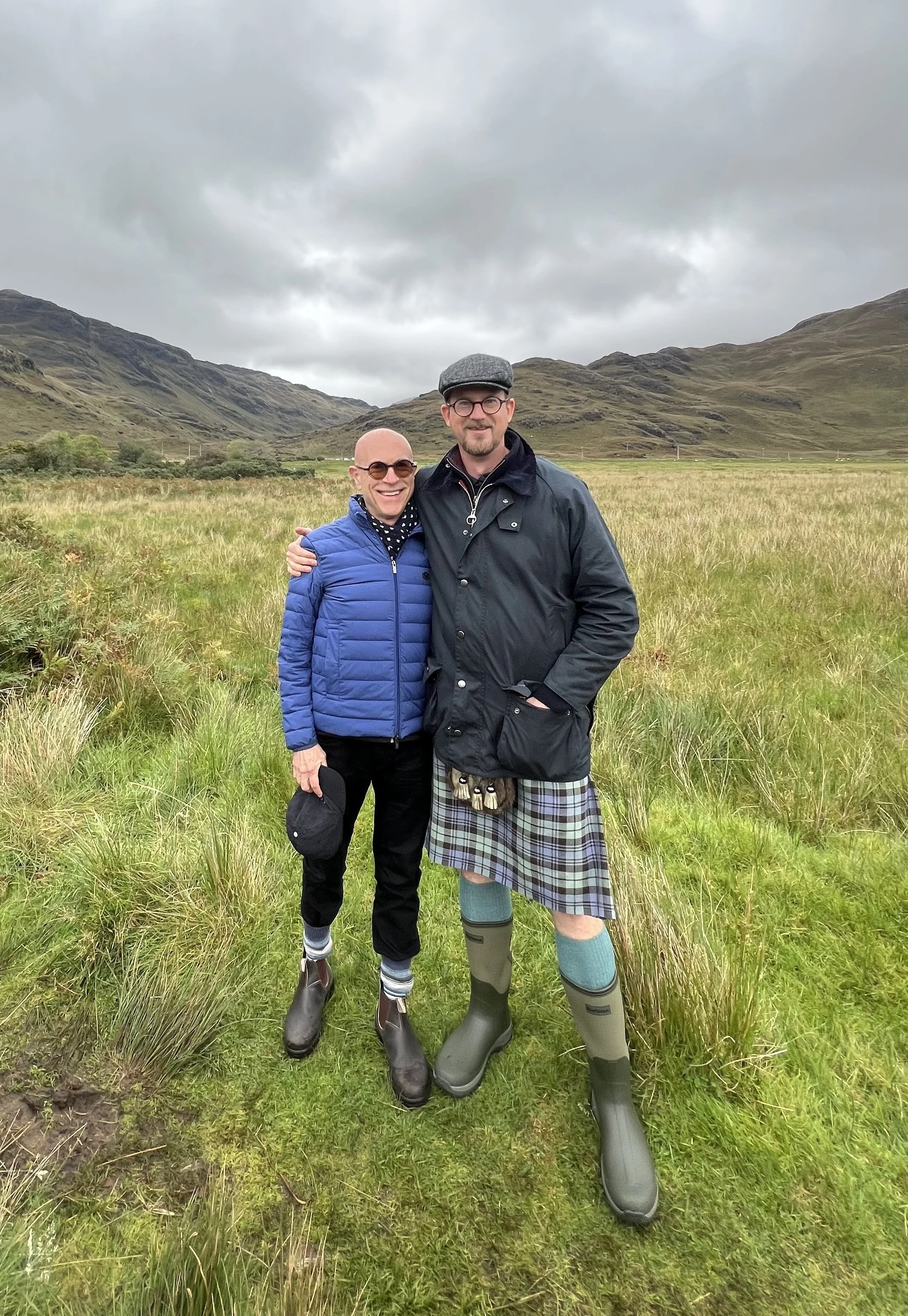 Two men standing together outdoors in a grassy field with mountainous terrain and cloudy sky in the background. They are wearing rain boots and jackets, one with a kilt.