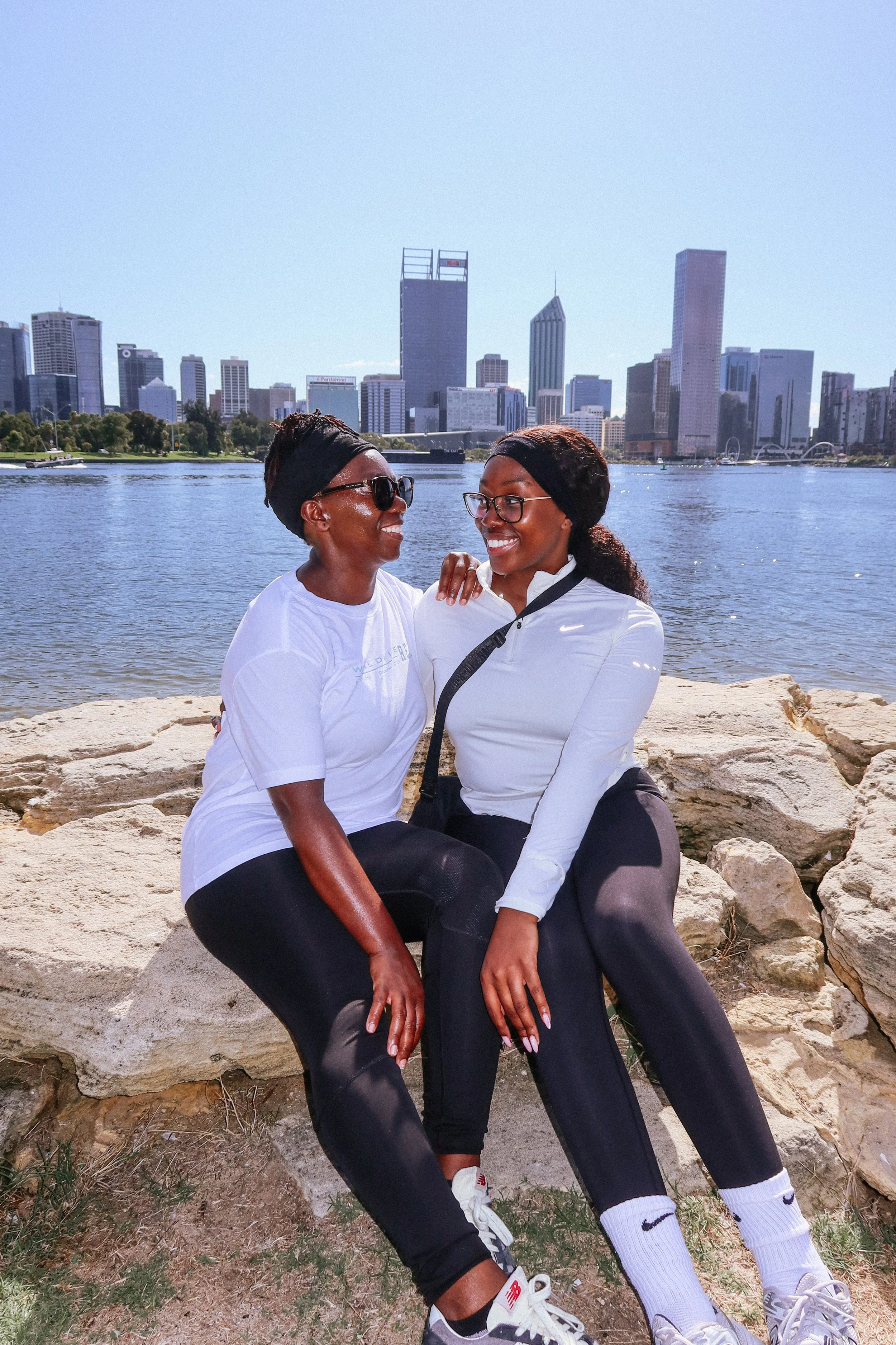 Two women sitting on rocks by a river with a city skyline in the background, smiling and looking at each other, wearing athletic clothing and headbands.