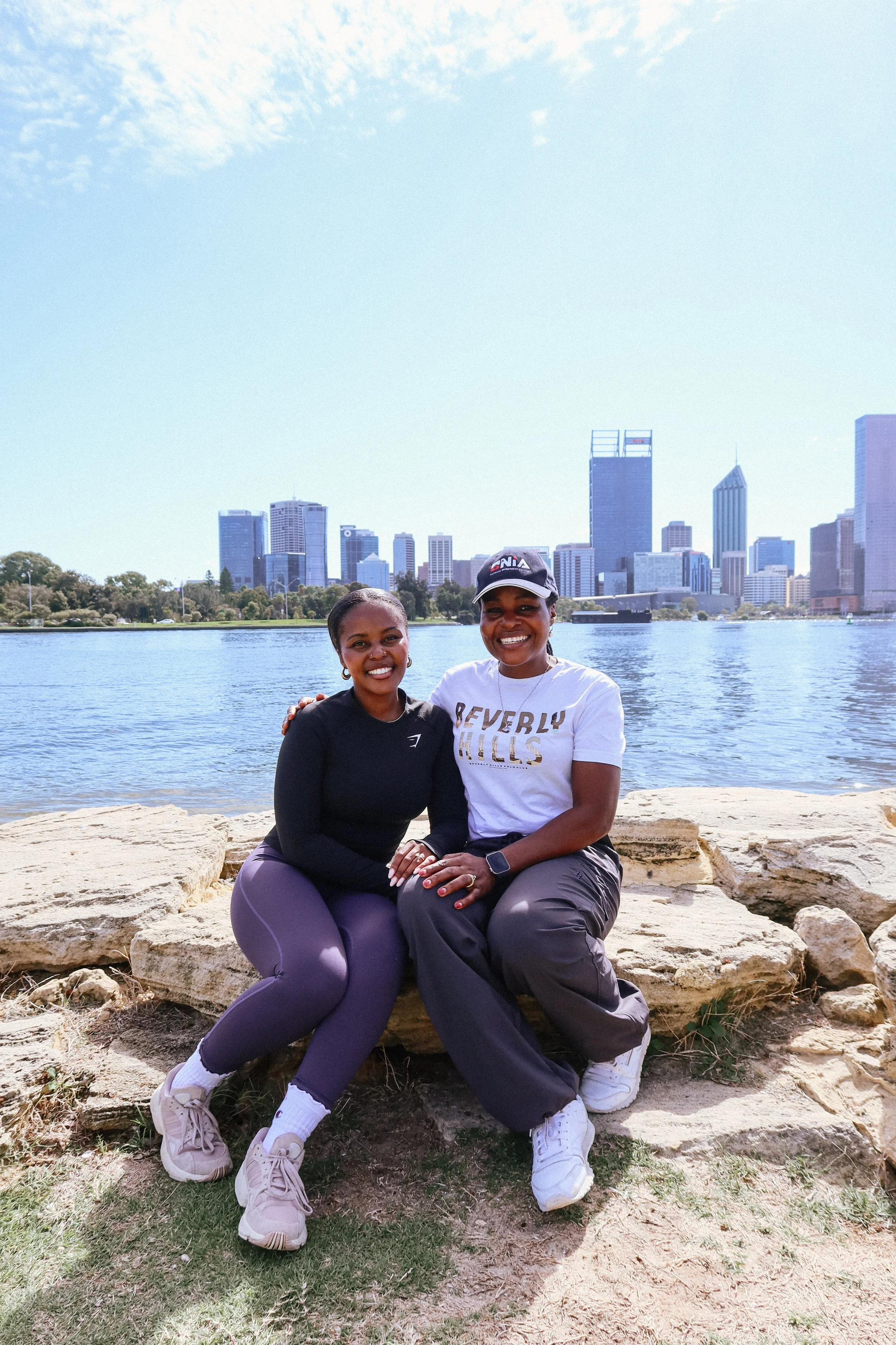 Two women sitting on a large rock by a body of water with a city skyline in the background. They are smiling, one wearing a black long-sleeve shirt and purple leggings, the other wearing a white T-shirt with text, gray pants, and a black cap. The sky