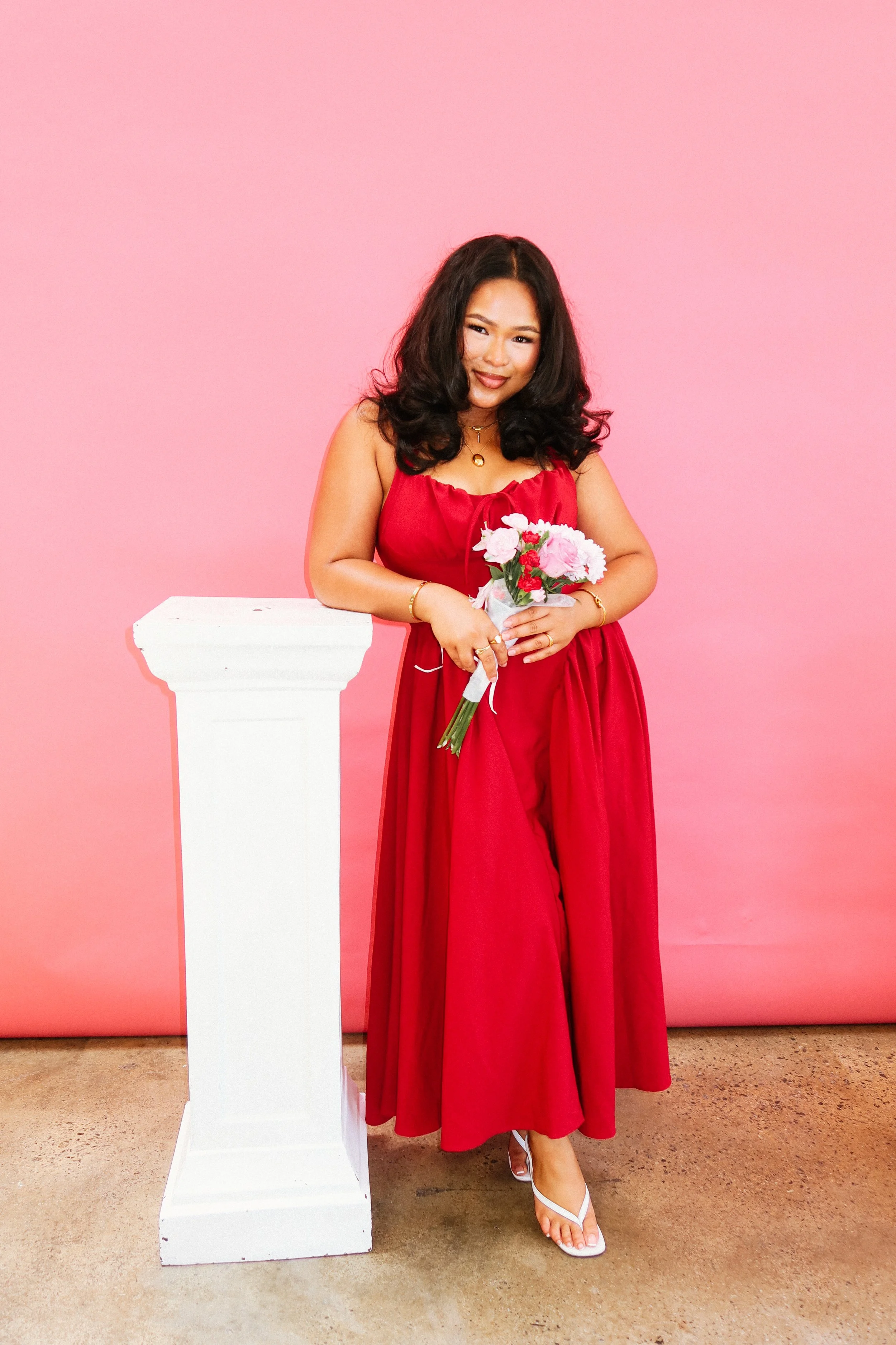 Woman standing next to a white pedestal against a pink background, at a Pink Dandelion community workshop, wearing a red dress, holding a bouquet of pink and white flowers.