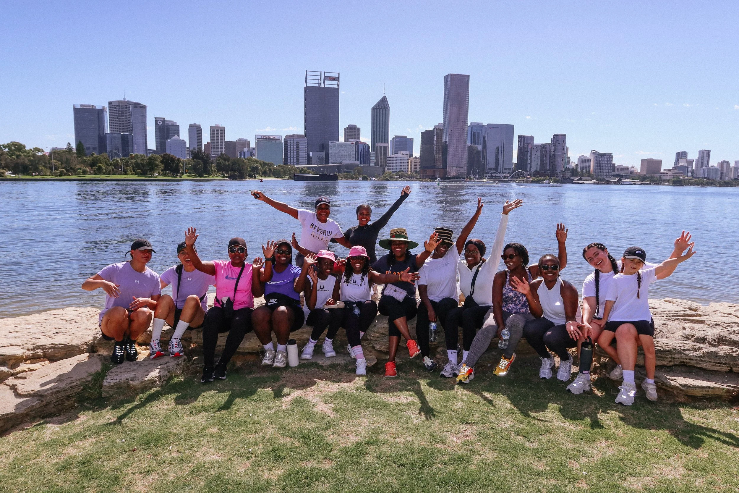Group of women and girls smiling and posing for a photo on a beach with a city skyline and water in the background.