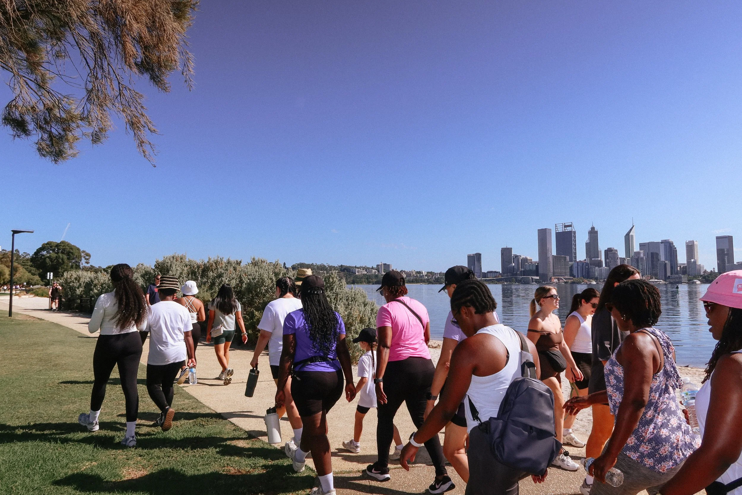 A group of women and girls walking along a path beside a river with a city skyline in the background and a clear blue sky.