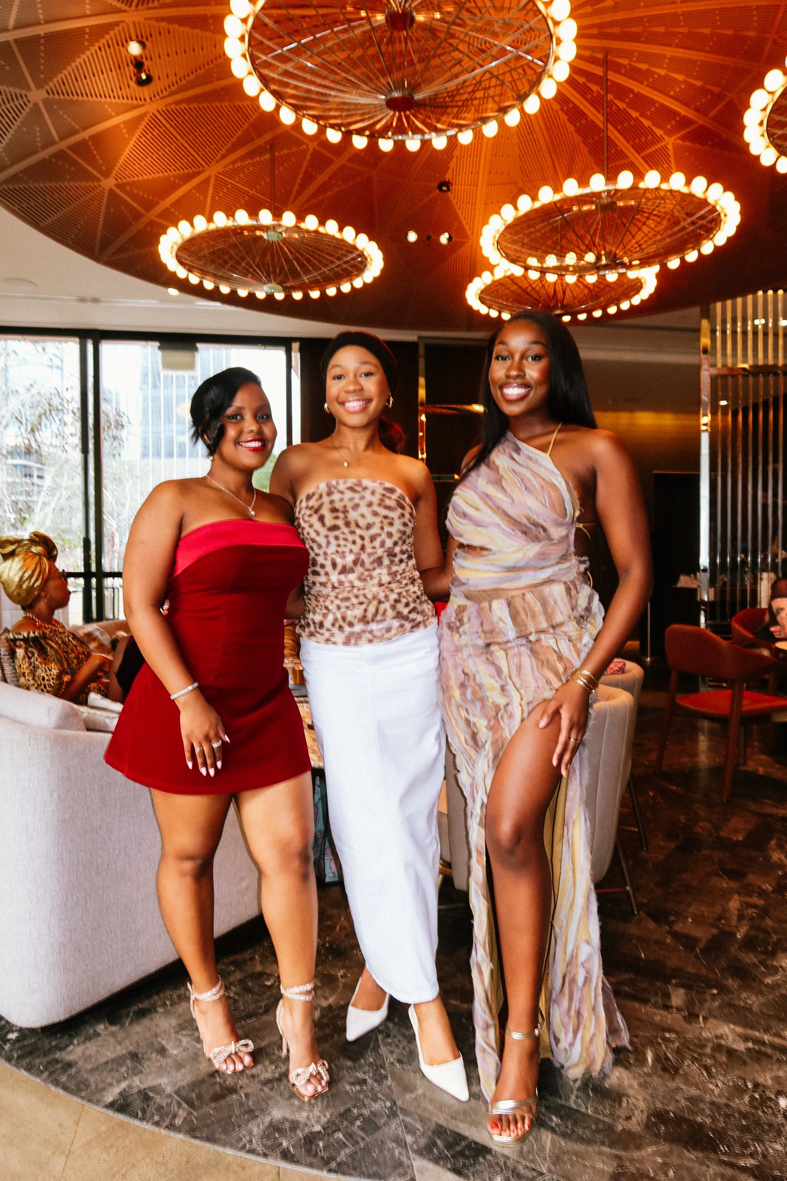 Three women standing together in a restaurant lounge area, at a Pink Dandelion community event, dressed in fashionable outfits, smiling at the camera.
