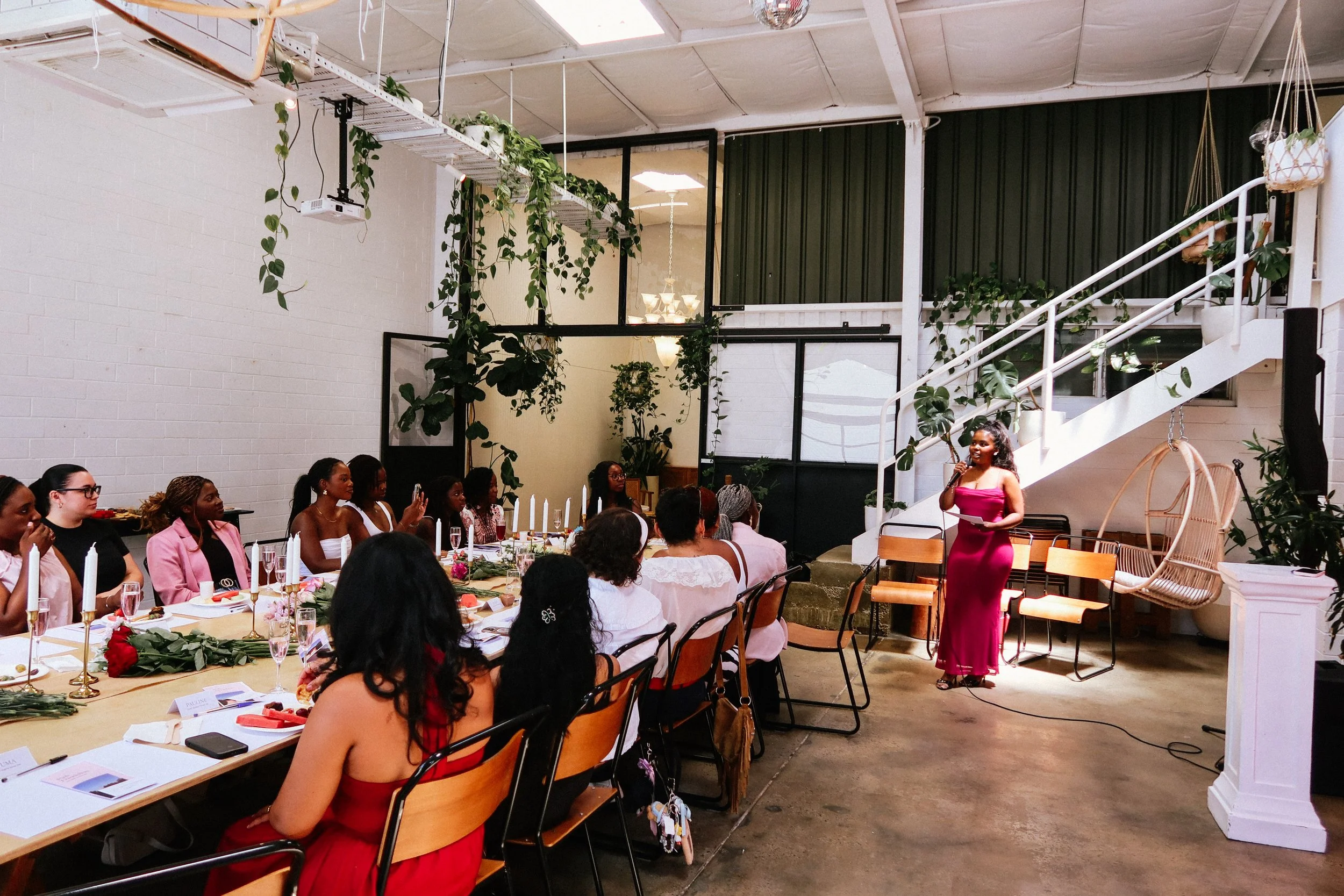 Women attending a Pink Dandelion workshop, seated around a decorated table with candles, and floral arrangements, while a woman in a pink dress speaks at a small stage in a modern, indoor venue with white walls, hanging plants, and large windows.