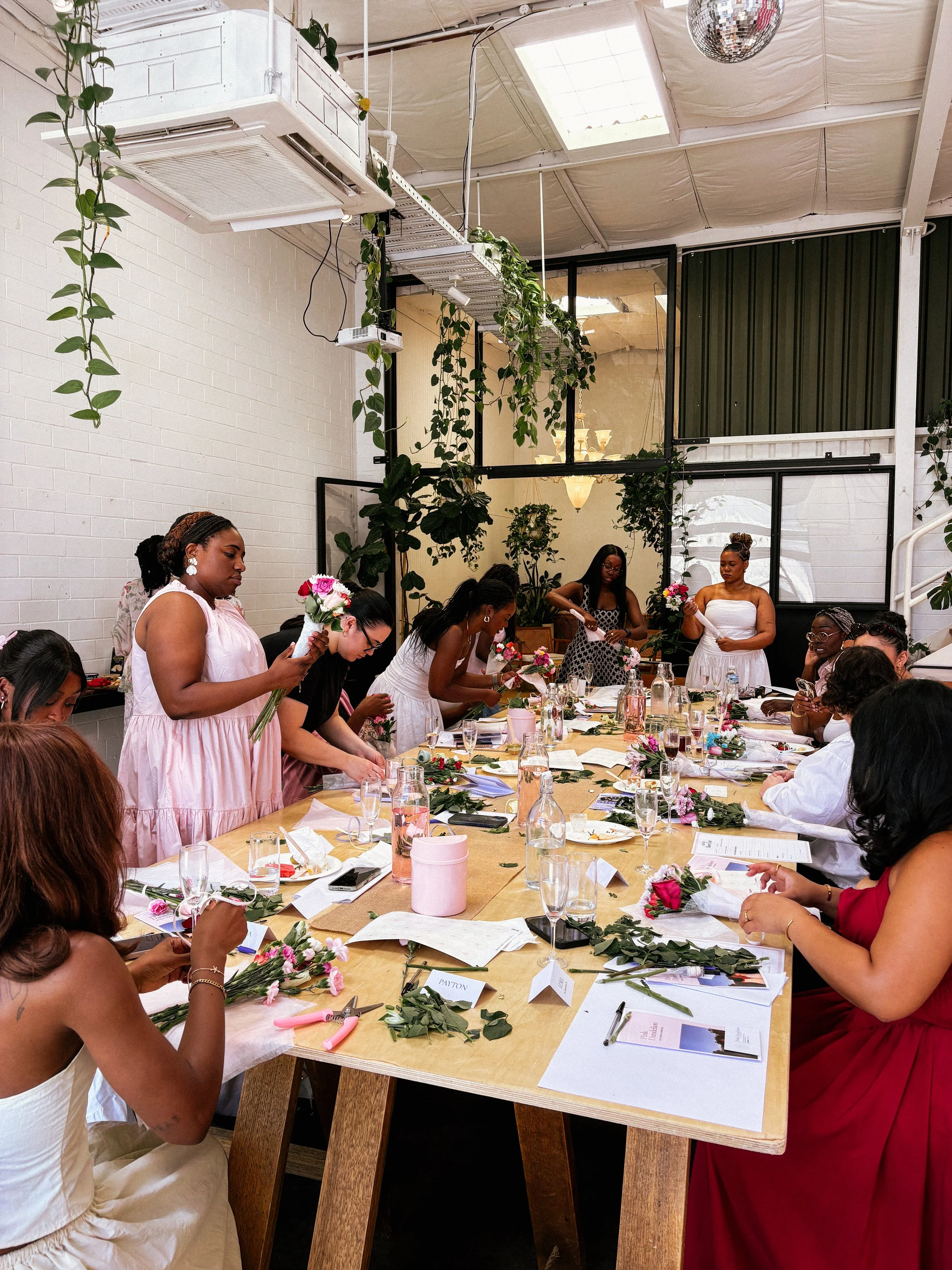 Women gathering around a long wooden table decorating flowers, at a Pink Dandelion community workshop, with greenery and pink blossoms, in a bright indoor space with white brick walls, plants, and large windows.