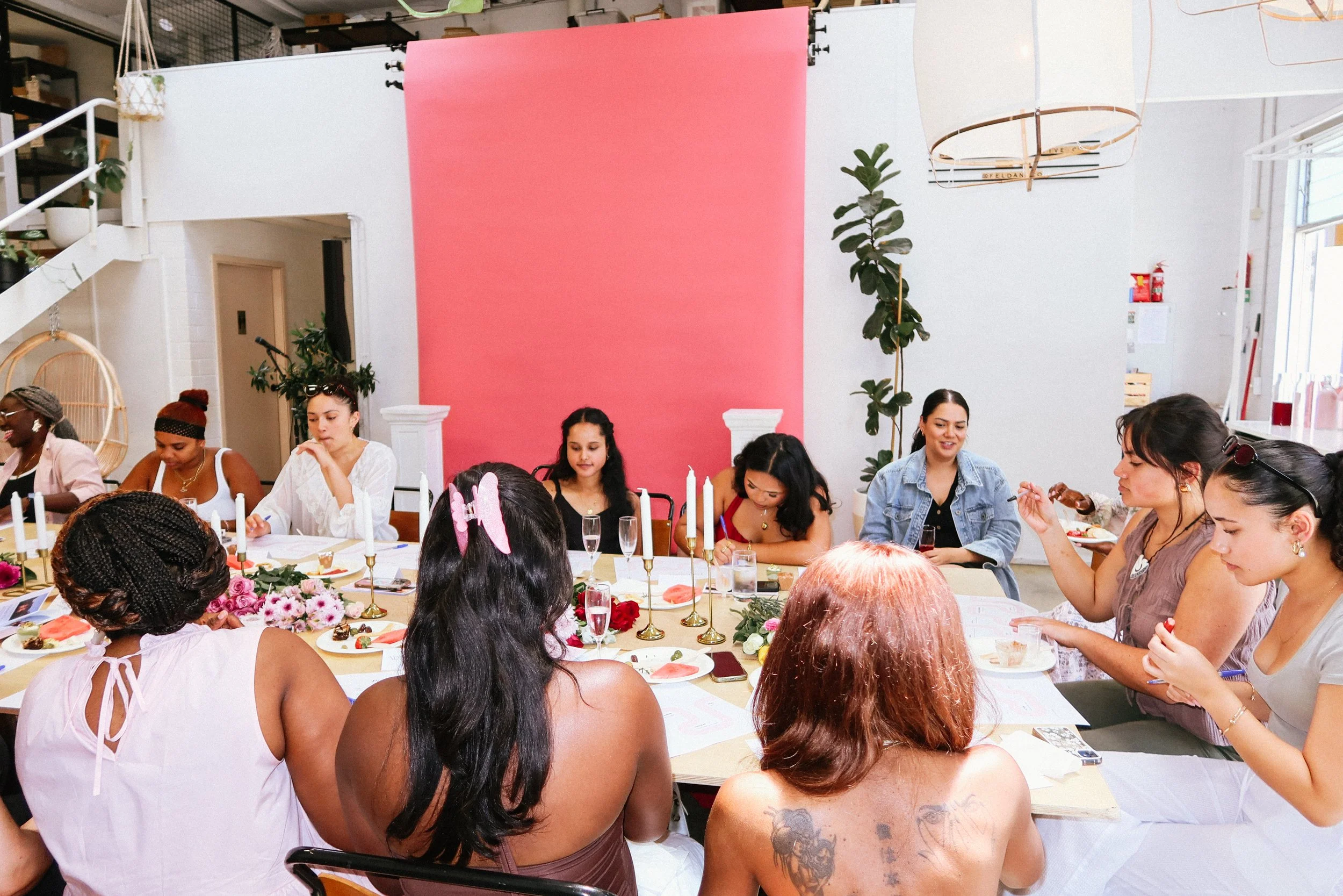 A diverse group of women sitting around a decorated table, at a Pink Dandelion community workshop, engaging in conversation, in a brightly lit room with pink backdrop and modern decor.