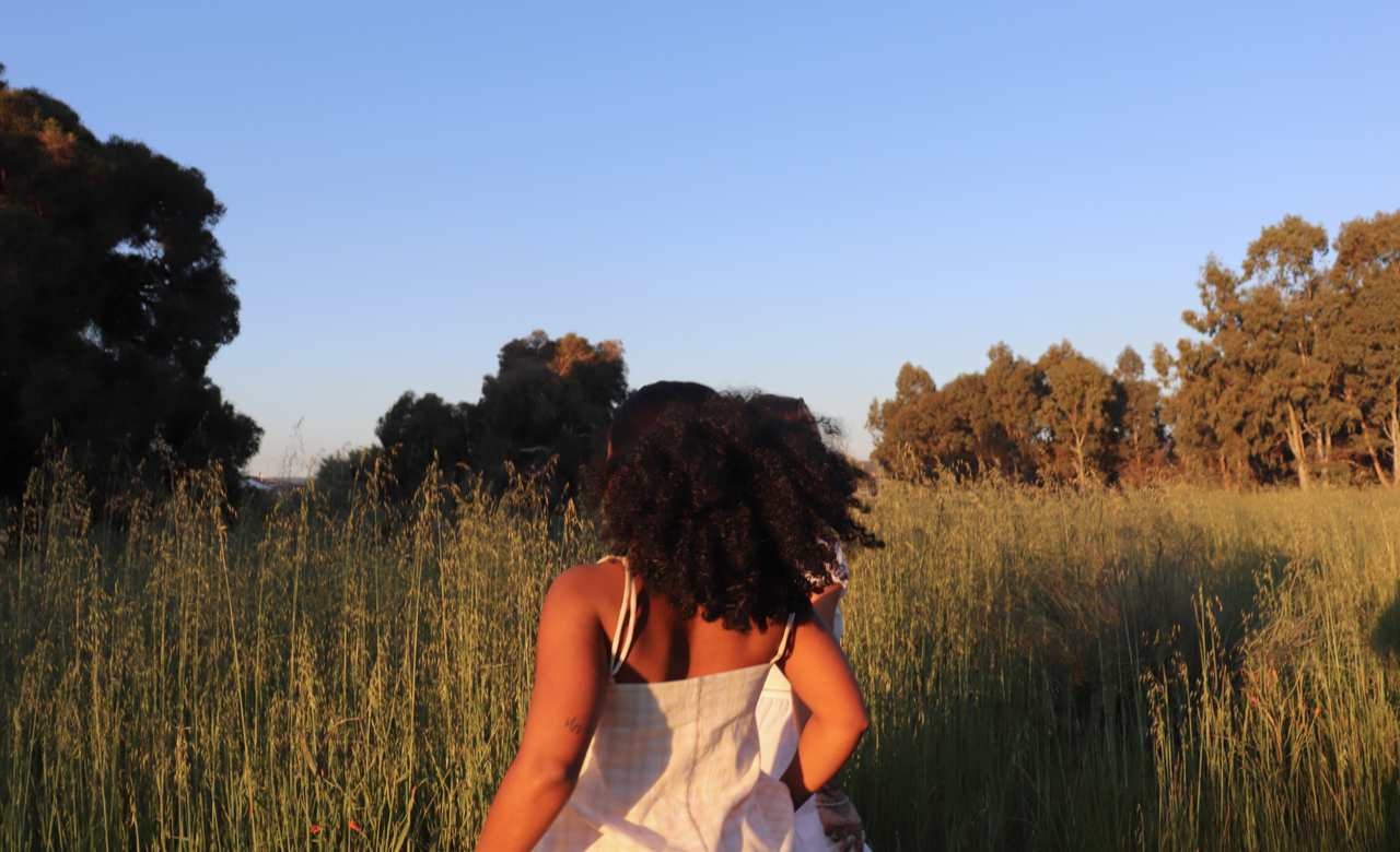 A woman with curly hair standing in a grassy field at sunset, facing away from the camera.