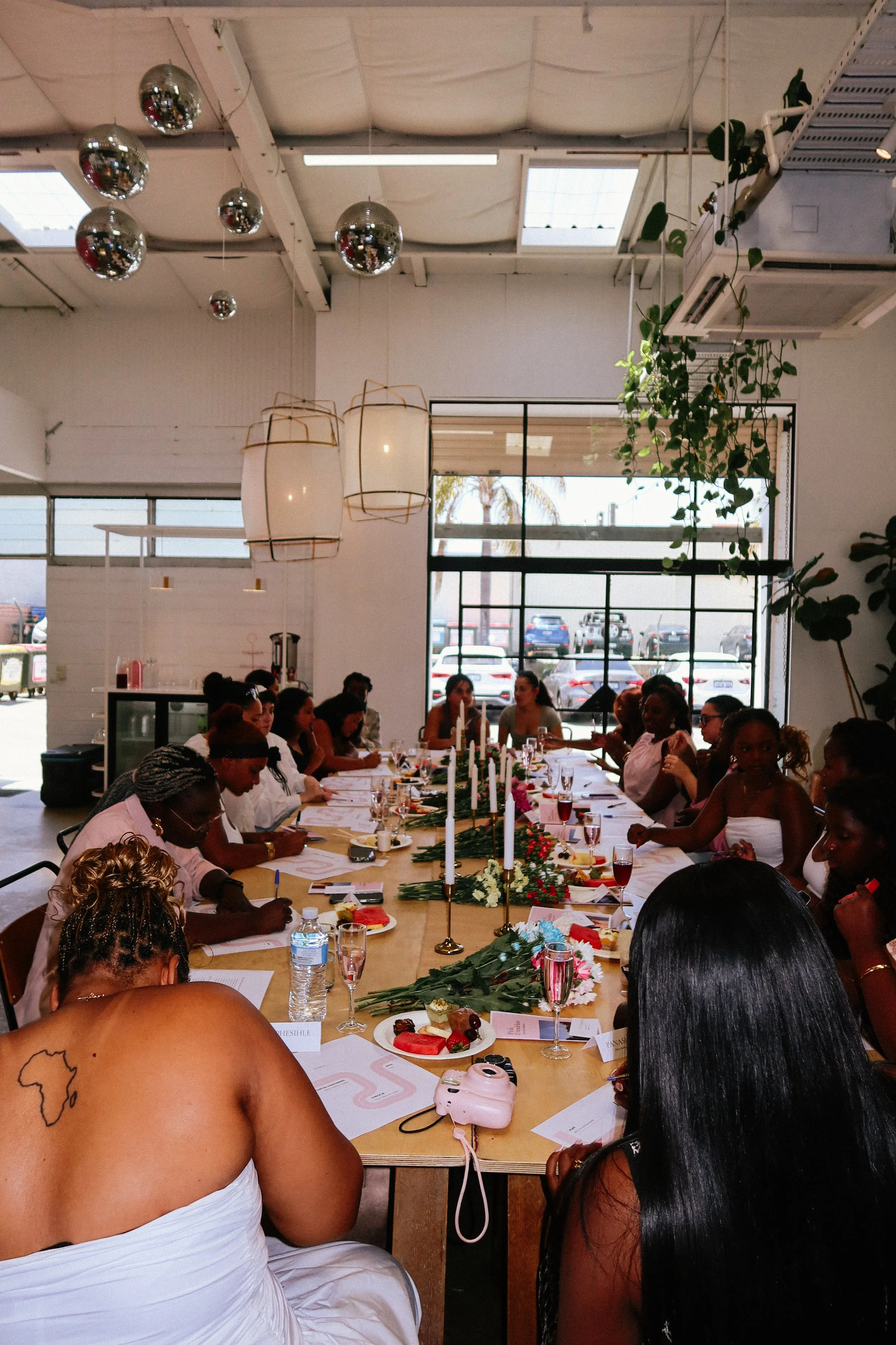 A large group of women at a Pink Dandelion community workshop, gathered around a long table in a bright, modern restaurant, with floral decorations and candles.