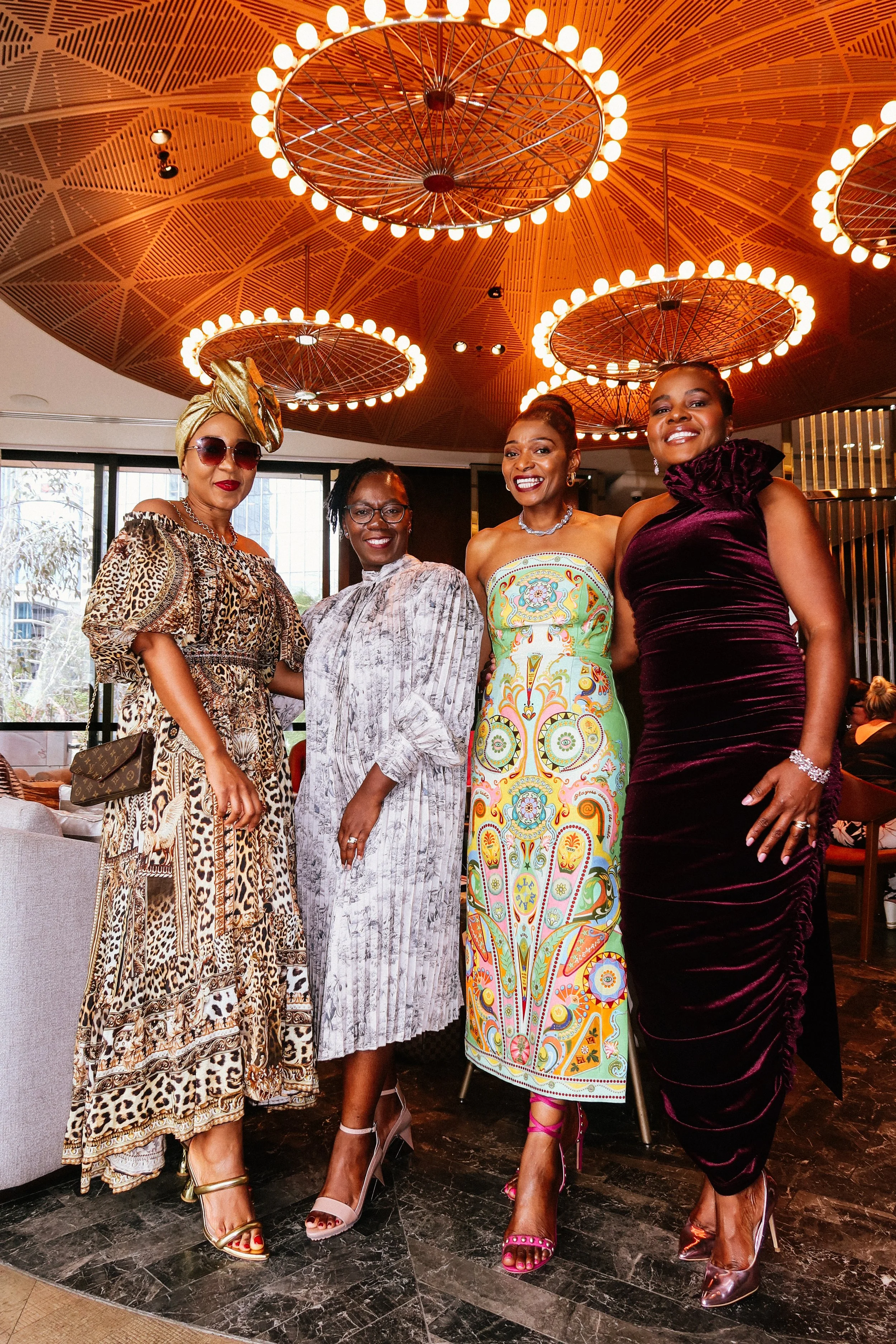 Four women dressed in fashionable and colorful attire standing together indoors, at a Pink Dandelion community event, smiling for a photo, with a modern, well-lit interior background.
