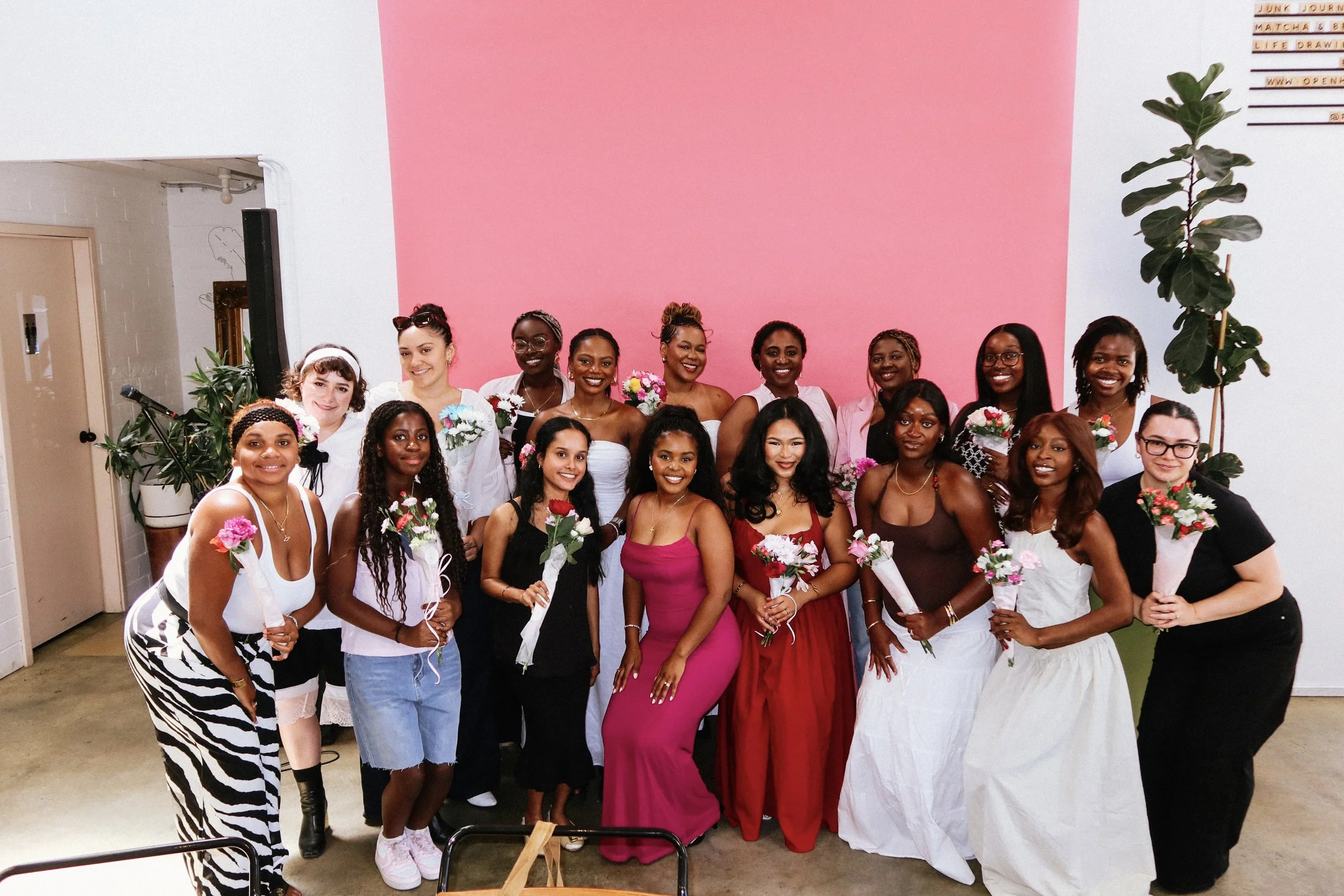 Group of women and girls posing indoors against a pink wall, at a Pink Dandelion community workshop some holding bouquets of flowers, dressed in various outfits including dresses, skirts, and casual wear, smiling at the camera.