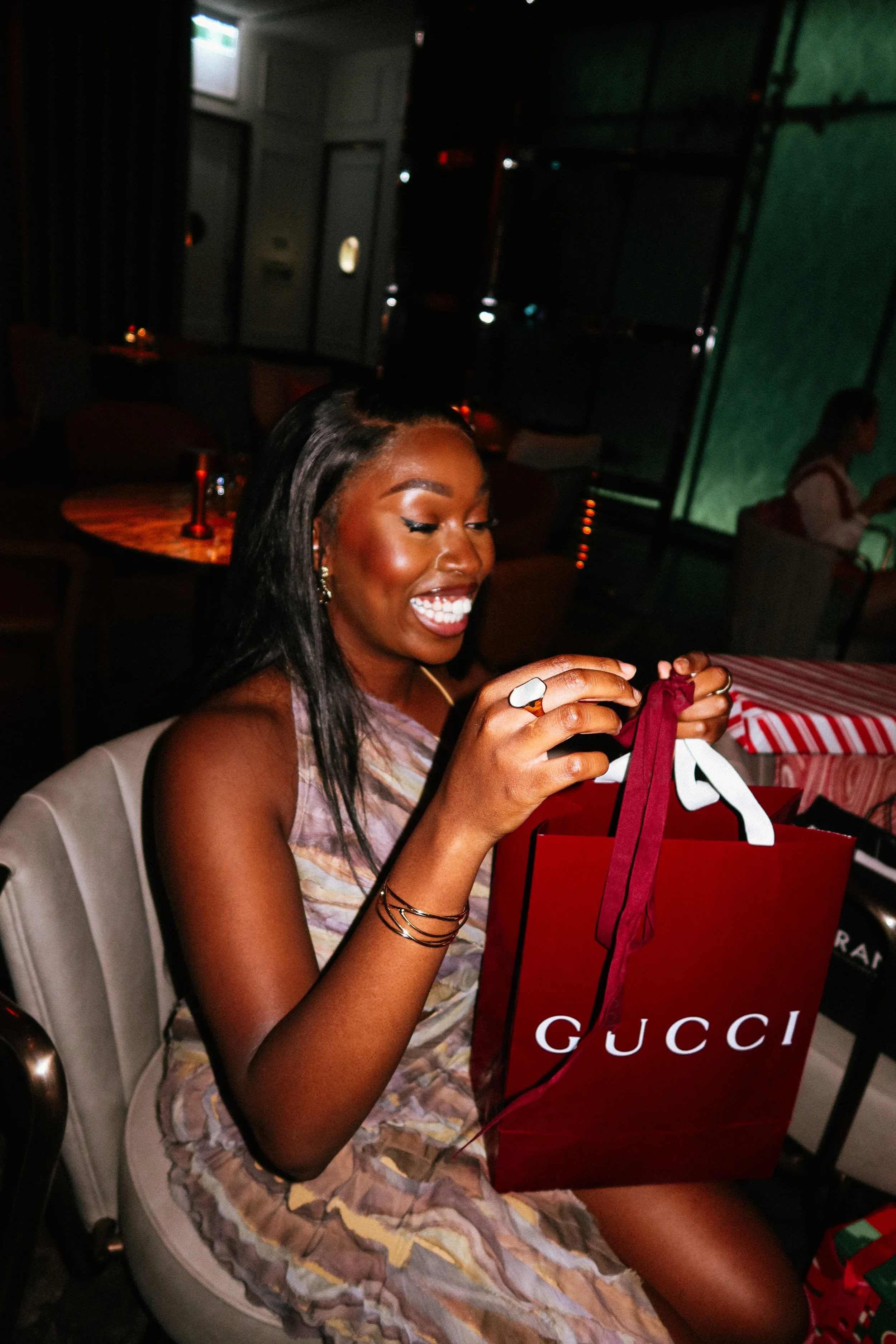 A woman with dark hair and a big smile sits at a table in a dark, stylish restaurant, opening a red Gucci shopping bag, at a Pink Dandelion community event.
