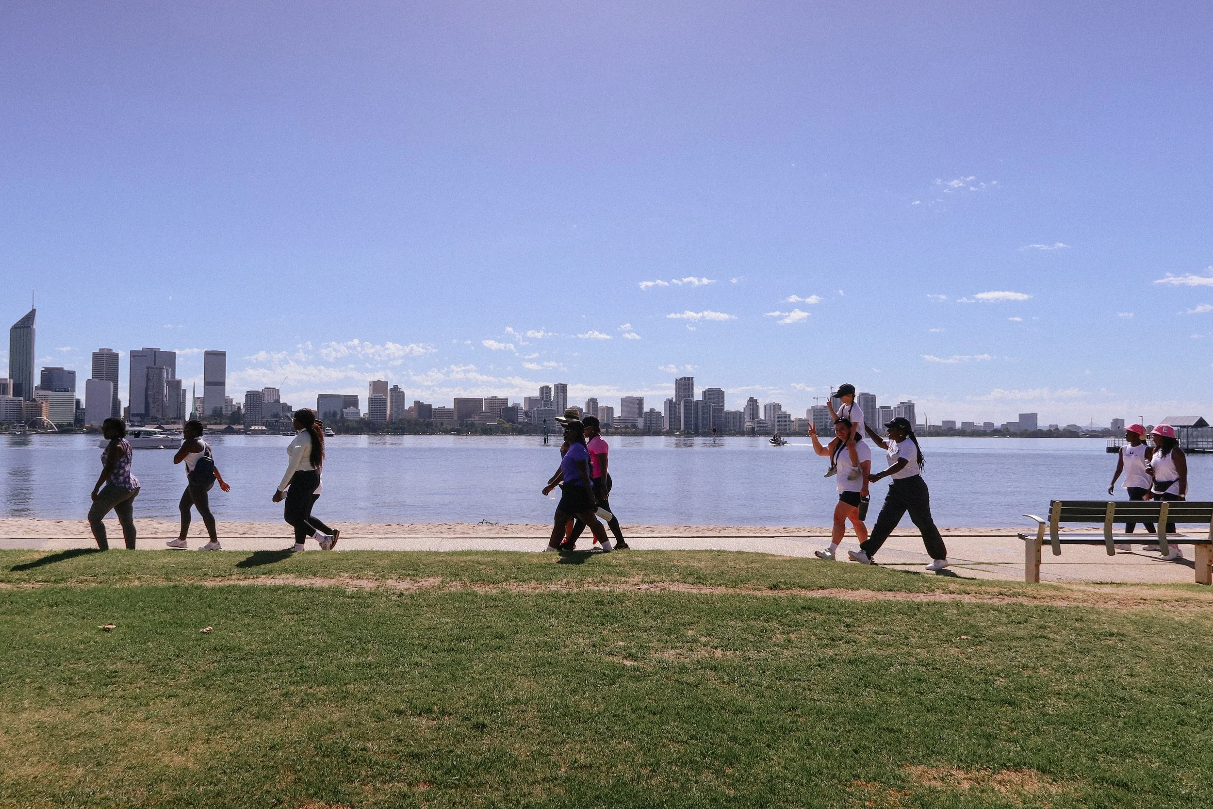 Women and girls walking along a pathway beside a body of water with a city skyline in the background. South Perth Foreshore. The sky is blue with some clouds.
