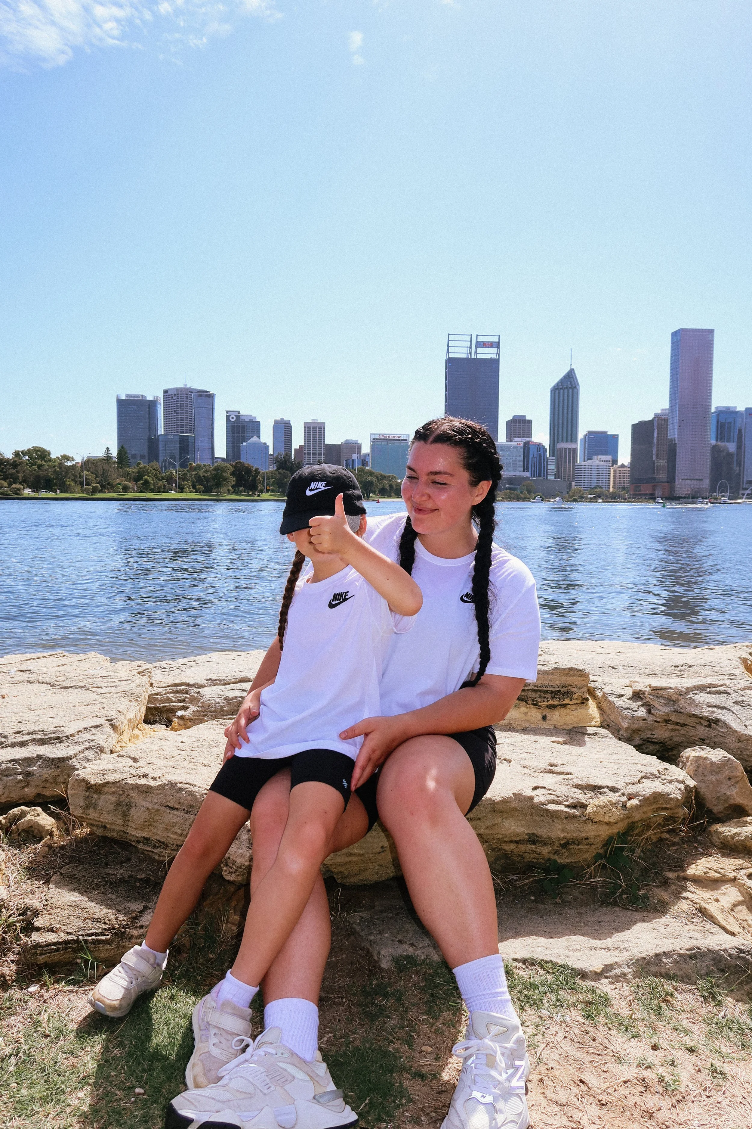 A woman and a child sitting on a large rock near a body of water with a city skyline in the background. The woman has dark braided hair and is giving a thumbs-up; the child is wearing a black Nike cap with a white swoosh, and they are both in white N