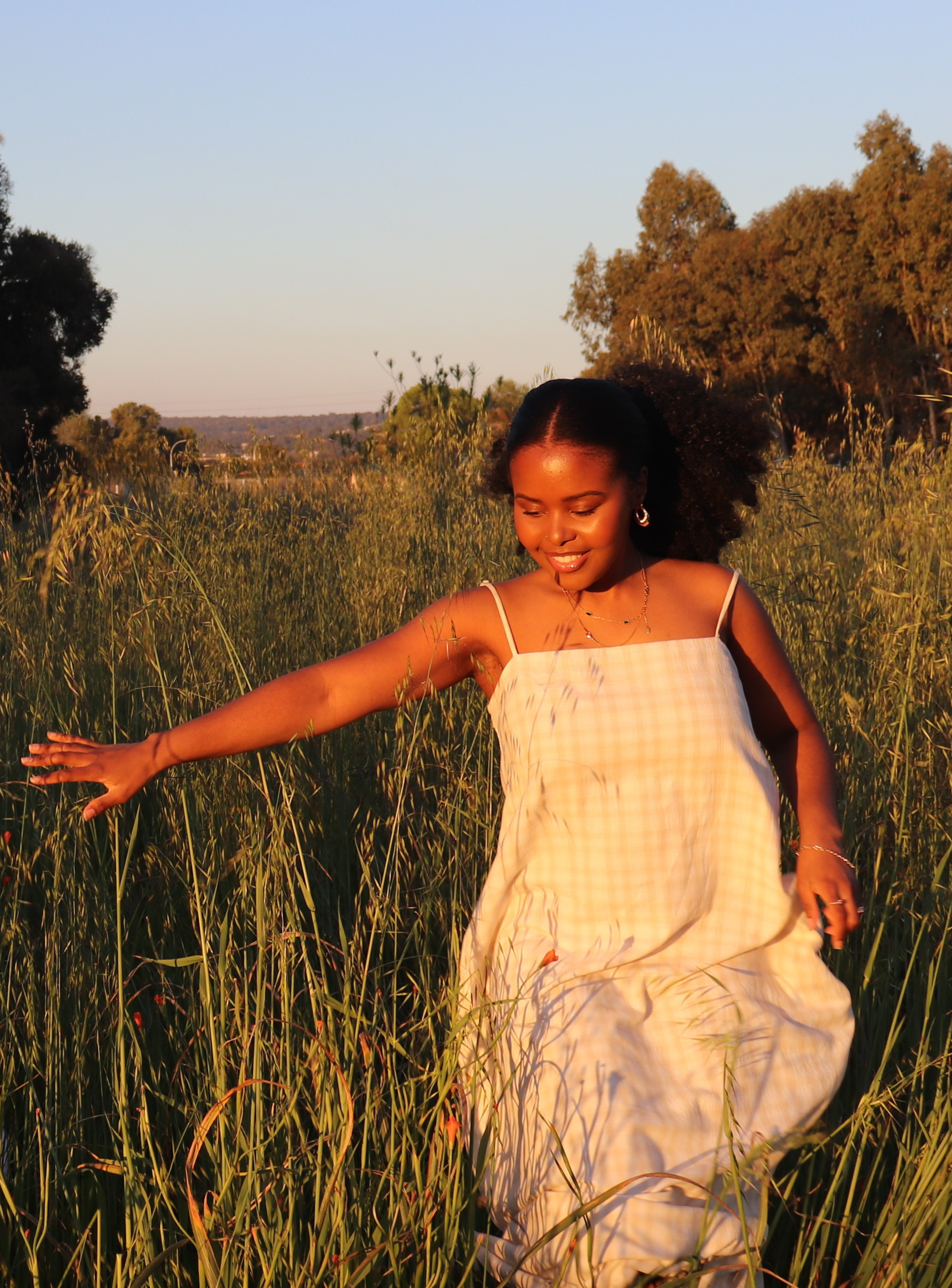 Woman walking through a field of long grass under blue skies, symbolising leadership, self-trust, clarity, and a grounded approach to wellness and mindset coaching.