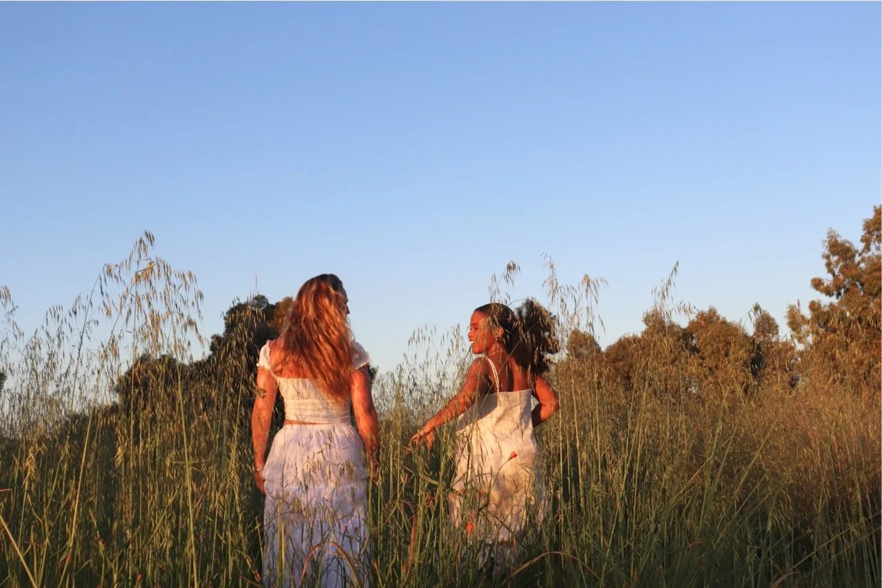 Three women in white dresses walking through a grassy field during sunset, smiling and engaging with each other.