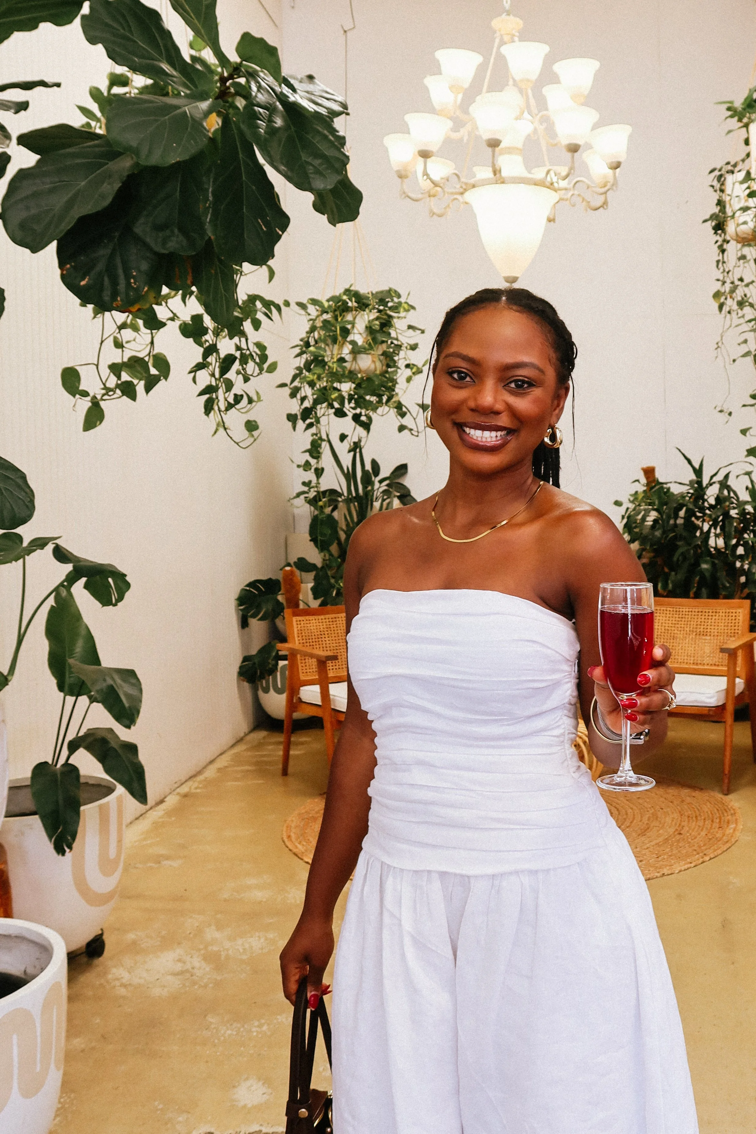 A woman in a white strapless dress holding a glass of red drink, at a Pink Dandelion community workshop, smiling in a room decorated with plants and a chandelier.