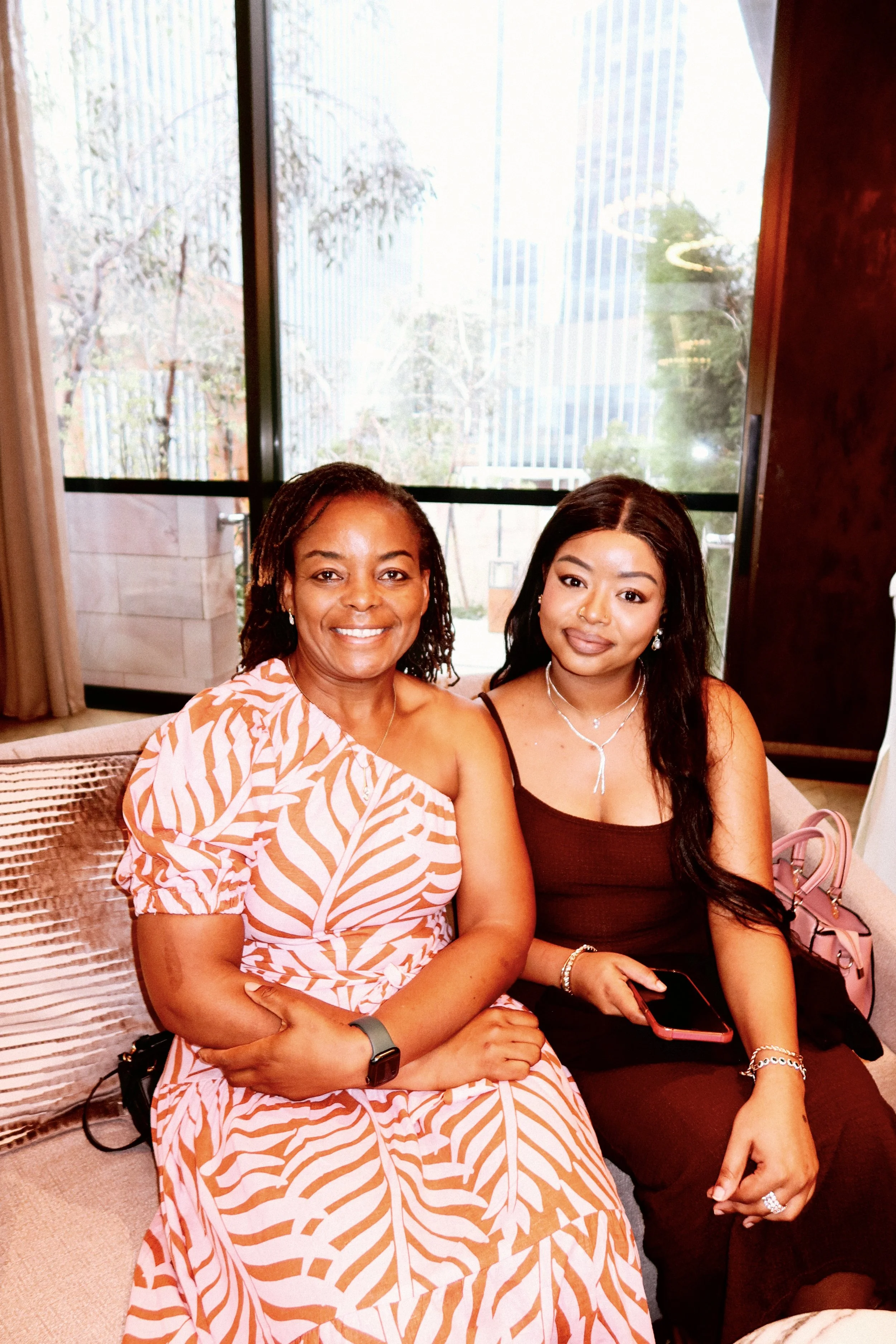 Two women sitting on a sofa in a well-lit room with large windows showing trees and buildings outside. The woman on the left is smiling, wearing a pink and white patterned dress, and the woman on the right is holding a phone, wearing a black tank top