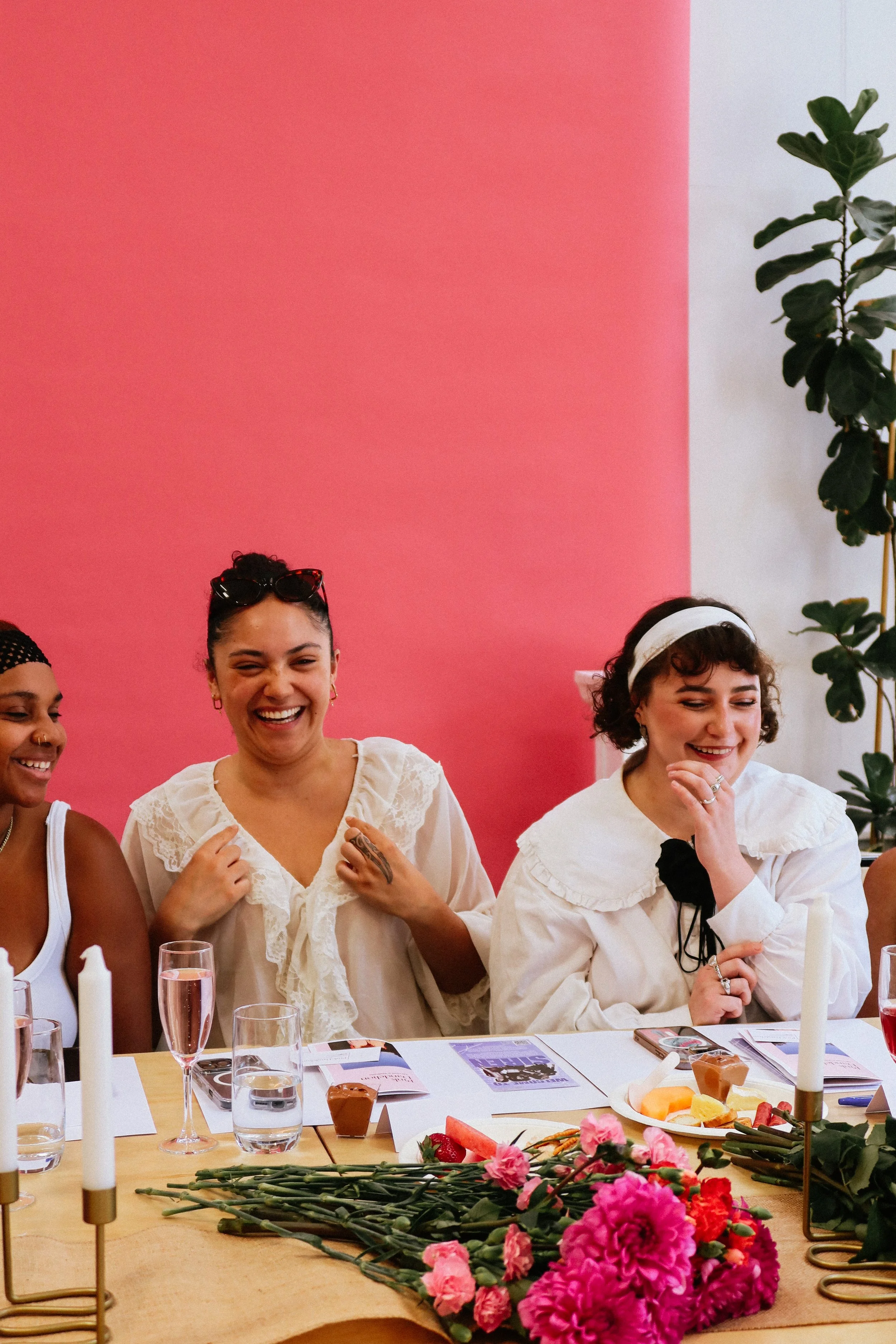 Three women sitting at a table at a Pink Dandelion community workshop, smiling and laughing, during a gathering or celebration in a room with pink and white walls, decorated with flowers and candles.