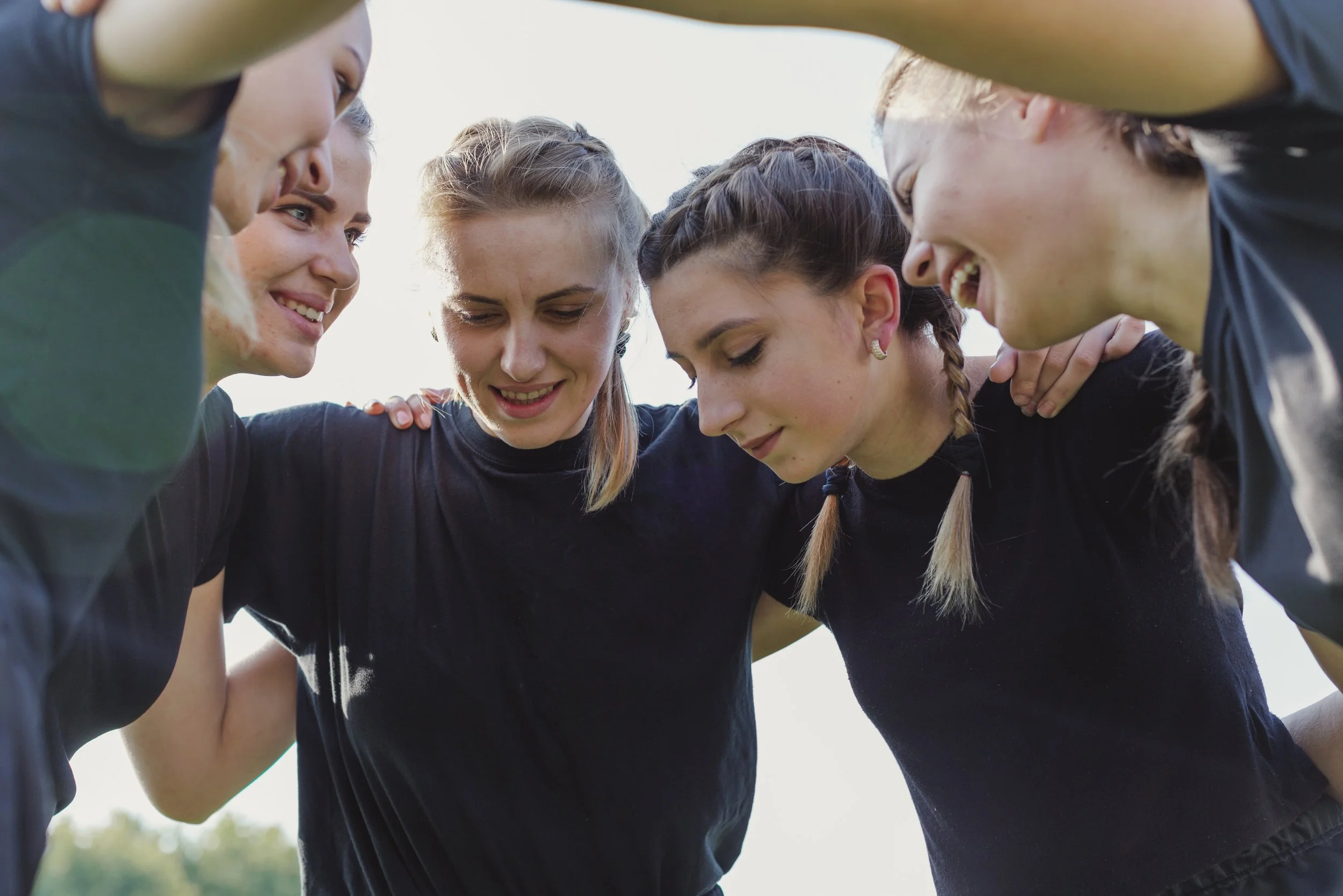 A group of women huddled together outdoors, showing unity and teamwork, with their heads bowed and arms around each other's shoulders.