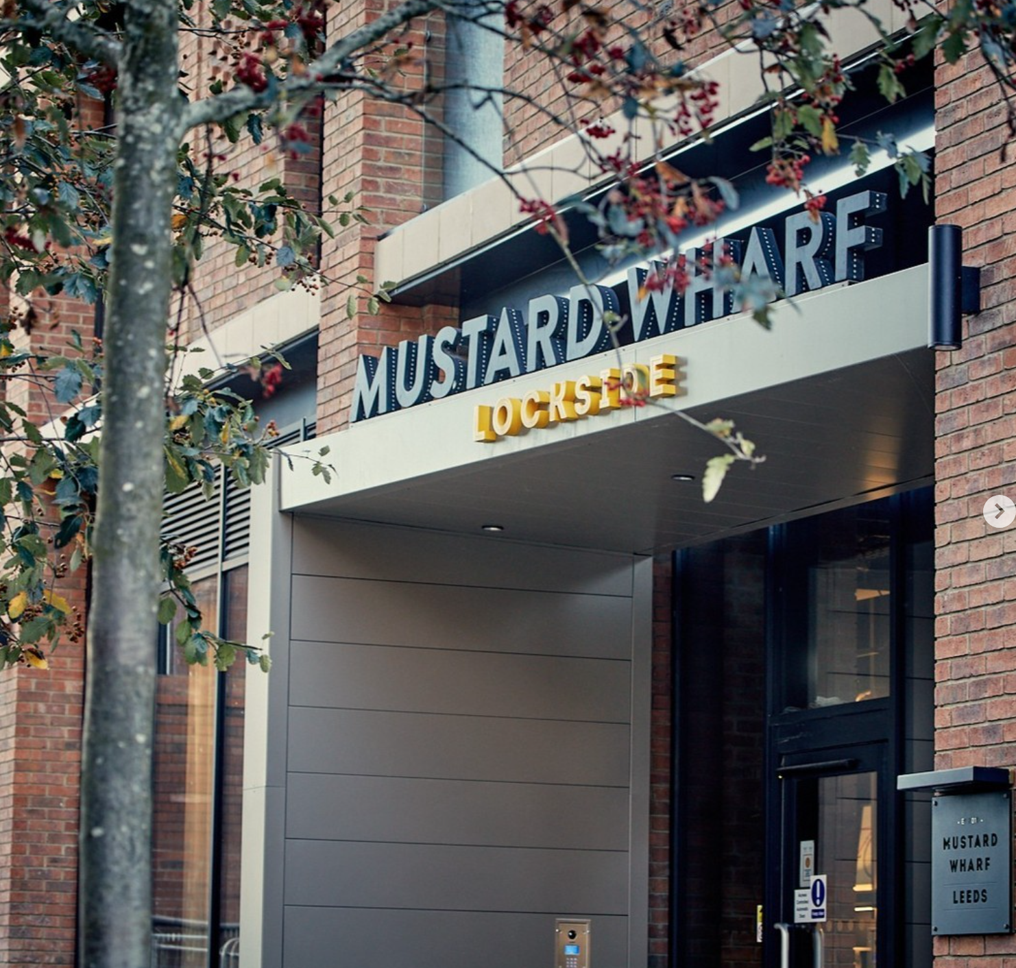 Exterior of a modern brick building with a sign that reads 'Mustard Wharf' and 'Lockside' beneath it. There are trees with red berries in the foreground.