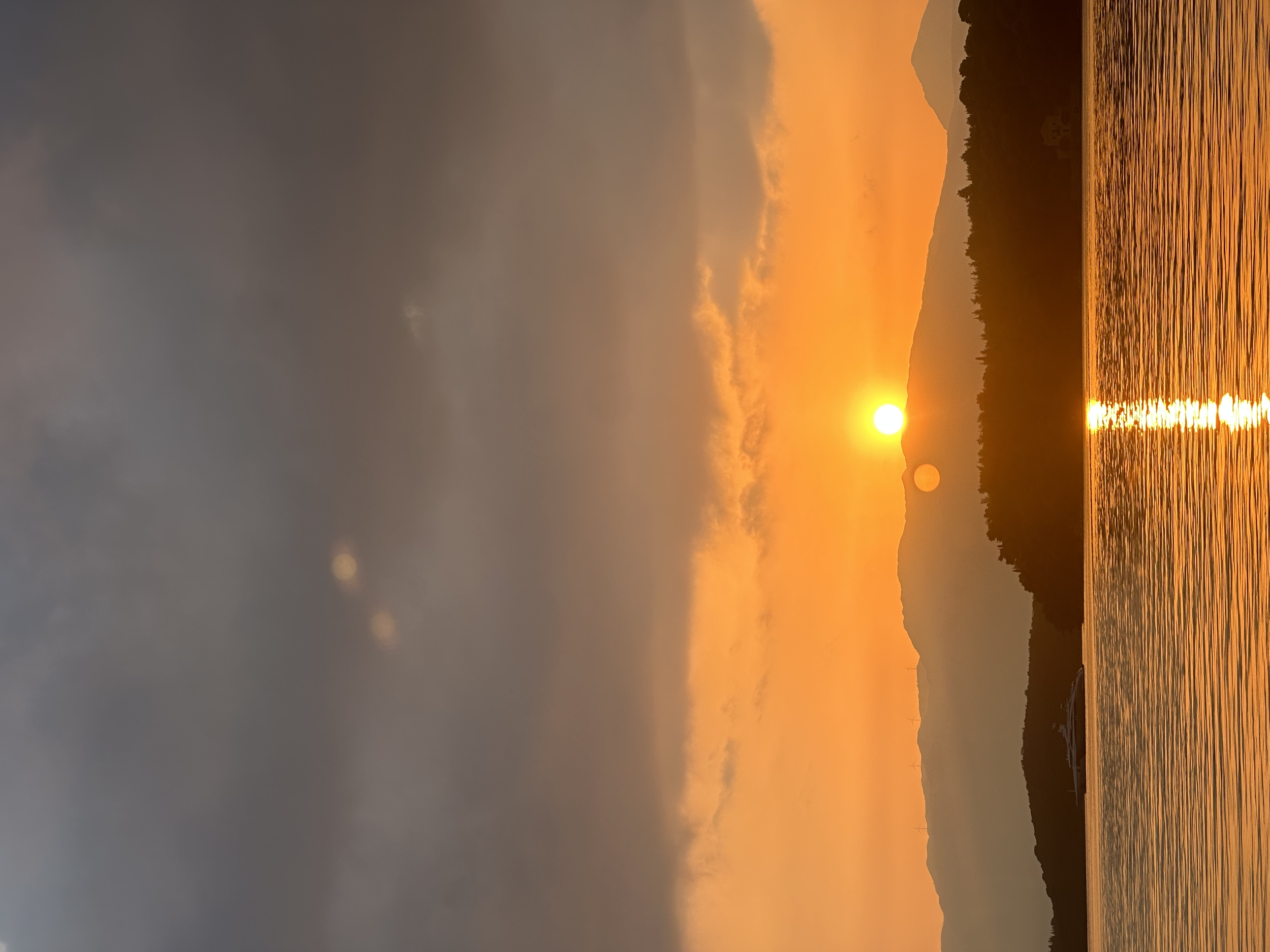 Sun setting over a body of water with mountains in the background and clouds in the sky.