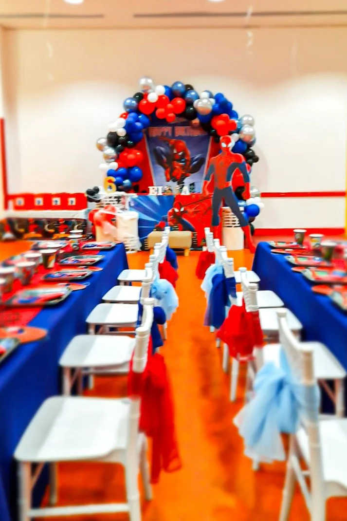 A birthday party table decorated with a Spider-Man theme. The table has blue tablecloths, red and blue decorations, and chairs with red and blue cloths. A backdrop with Spider-Man images and a large balloon arch in red, white, black, grey, and blue b