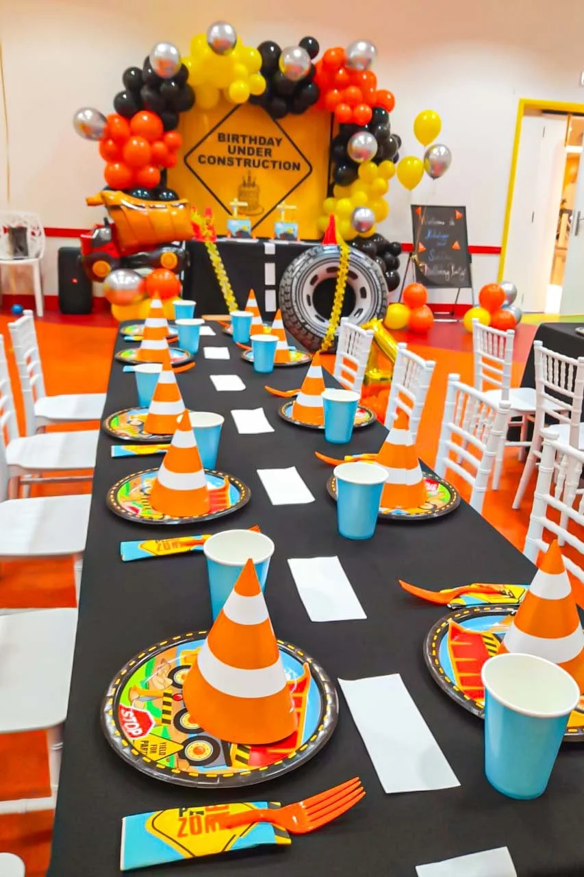 Colorful birthday party table with traffic cone decorations, blue cups, and race car themed plates. In the background, a balloon arch, a "Birthday Under Construction" sign, and construction-themed decorations including a toy dump truck, a tire, and c