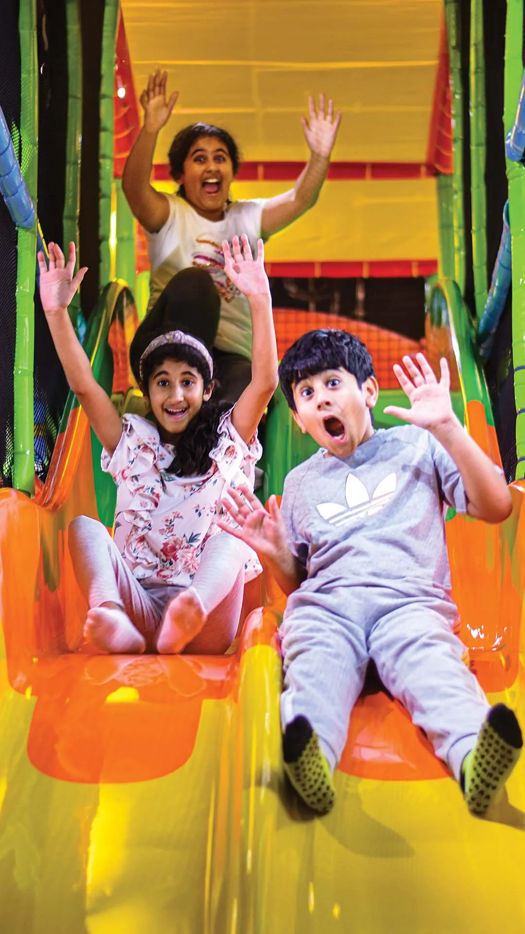 Four children sliding down an indoor colorful play structure, smiling and waving, with bright yellow, green, and orange colors.