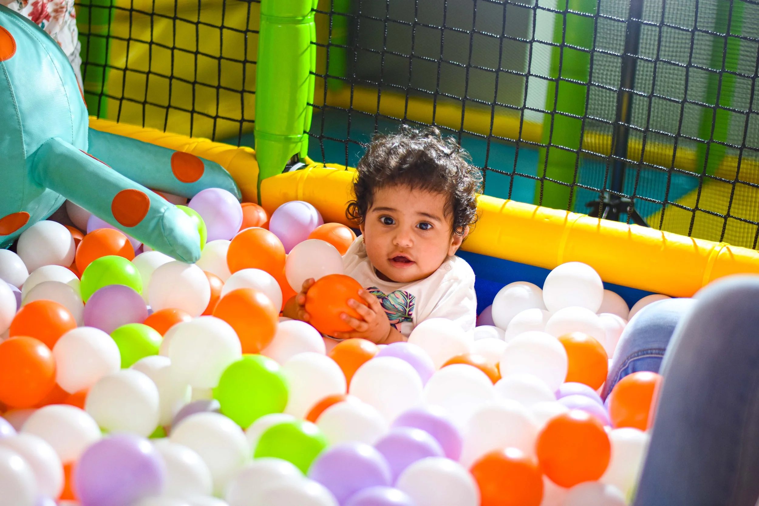 Young child with curly hair sitting in a colorful ball pit, holding an orange ball, surrounded by white, purple, orange, and green balls, with a netted play area in the background.