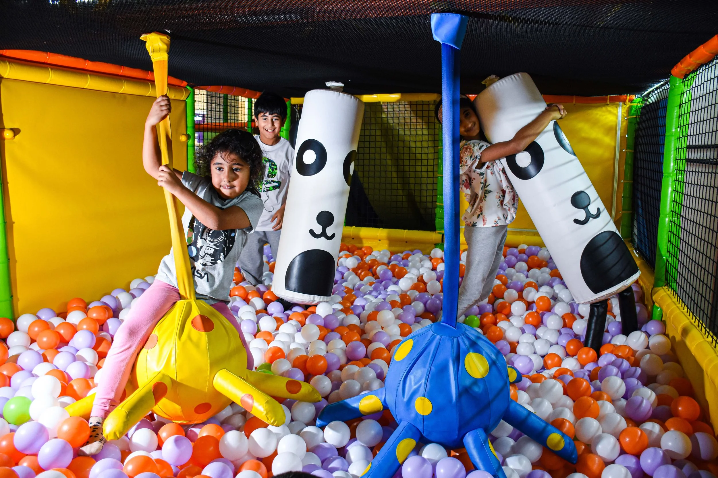Children playing in a colorful ball pit with plush octopus toys and foam dog-shaped cushions.