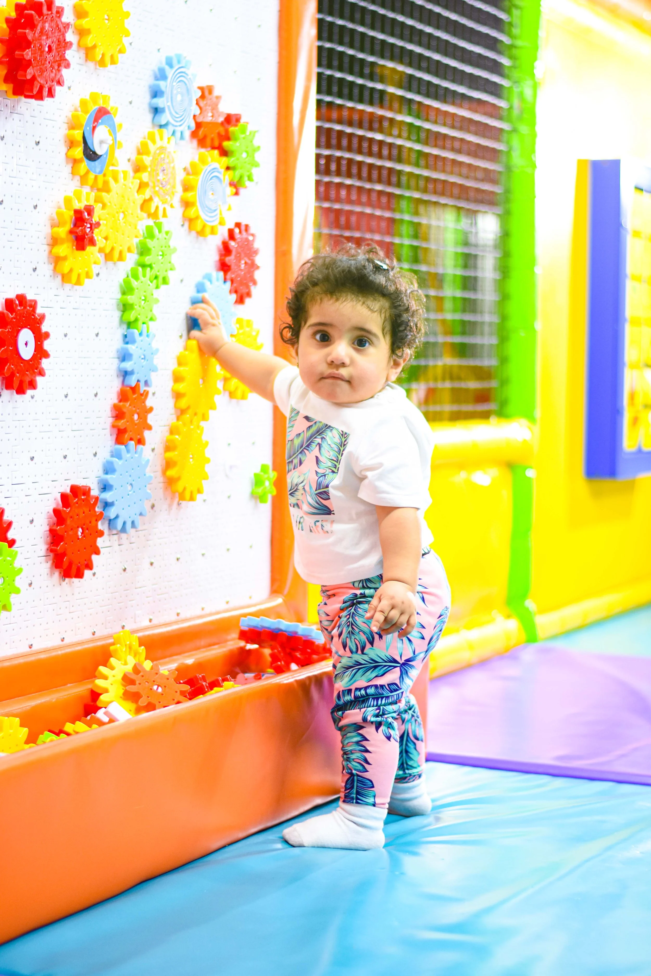 Young child playing with colorful gear-shaped building blocks on a white pegboard wall in an indoor play area.