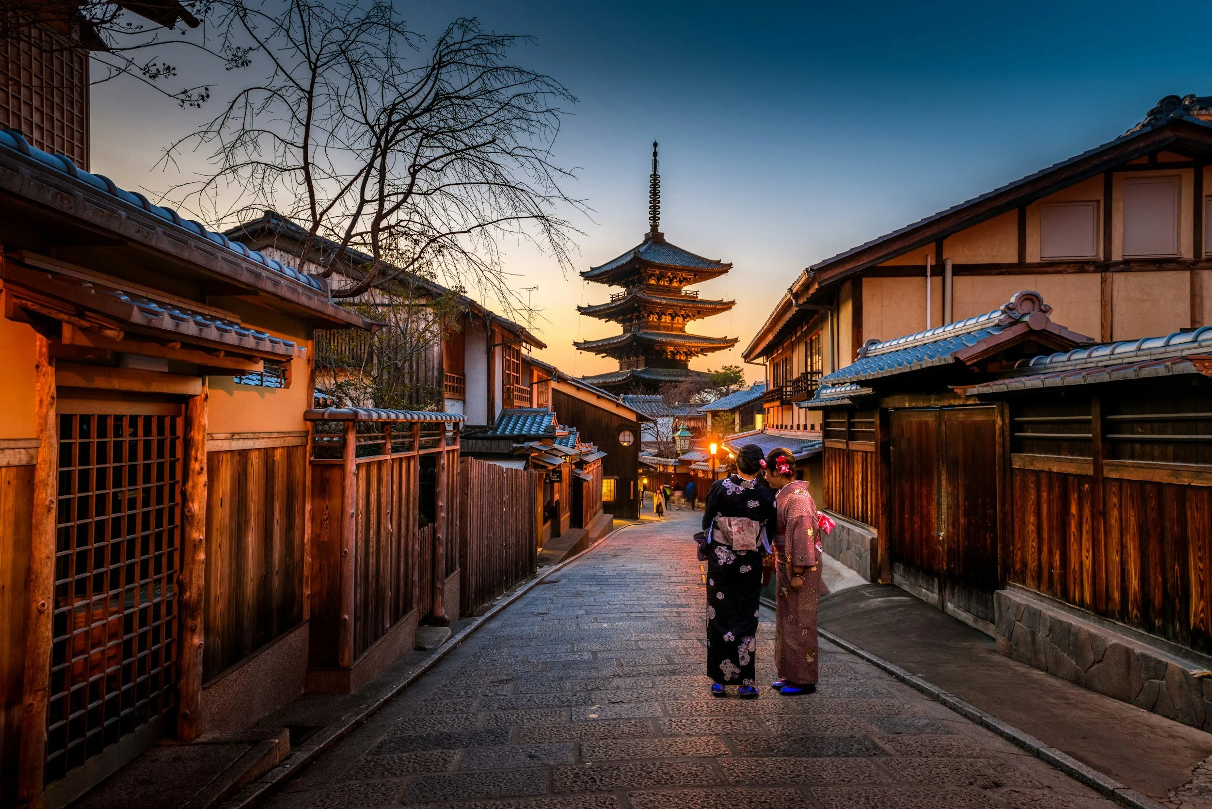 Deux femmes en kimono discutent dans une rue pavée traditionnelle japonaise, avec un temple pagode en arrière-plan au coucher du soleil.